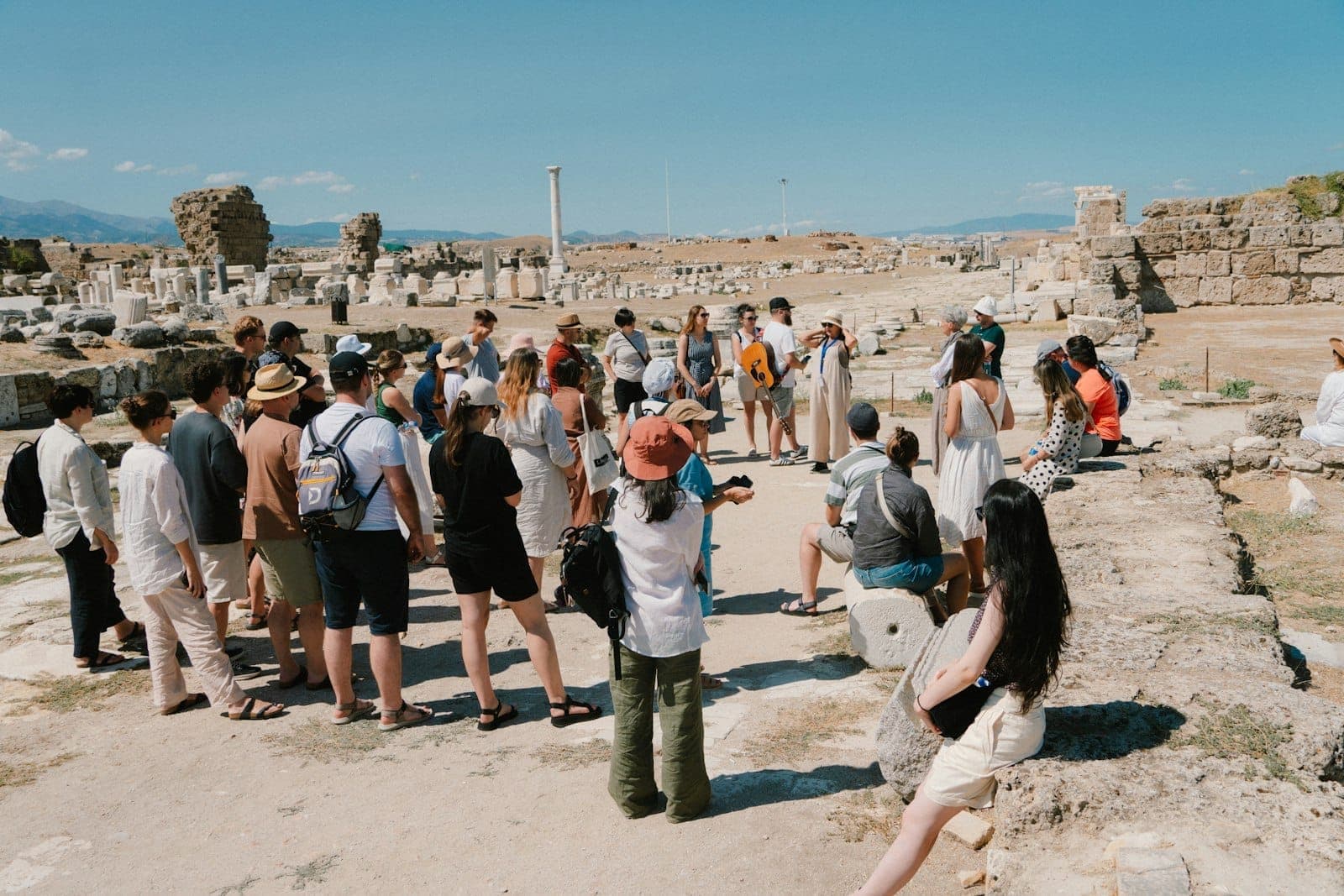Eine Gruppe von Touristen versammelt sich um einen Reiseführer inmitten antiker Ruinen und Steinsäulen unter klarem blauen Himmel