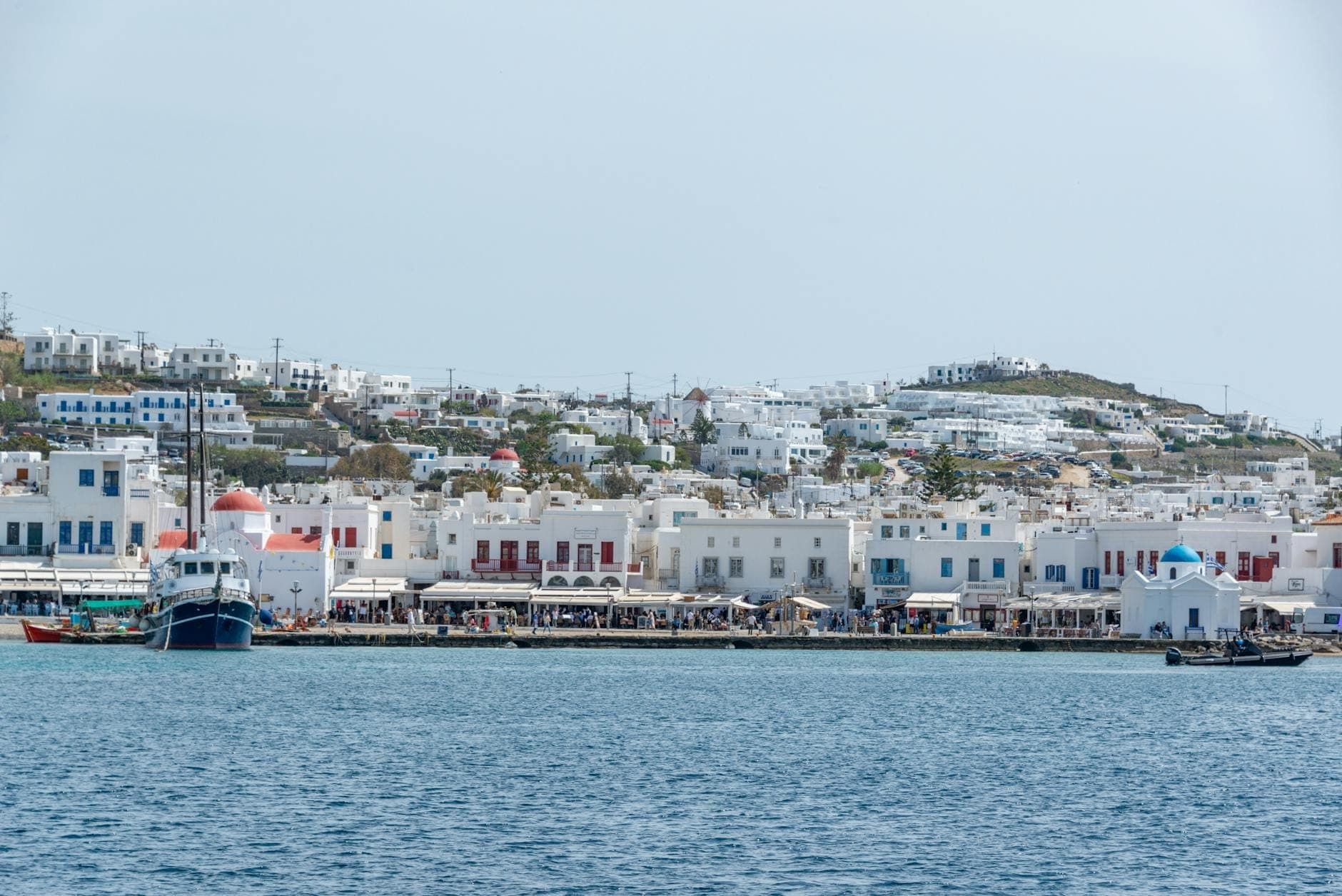 Blick auf die Uferpromenade von Mykonos mit angedockten Booten, weiß getünchten Gebäuden und der blaukuppligen Kirche St. Nicholas in der Nähe des Abfahrtspunkts nach Delos.