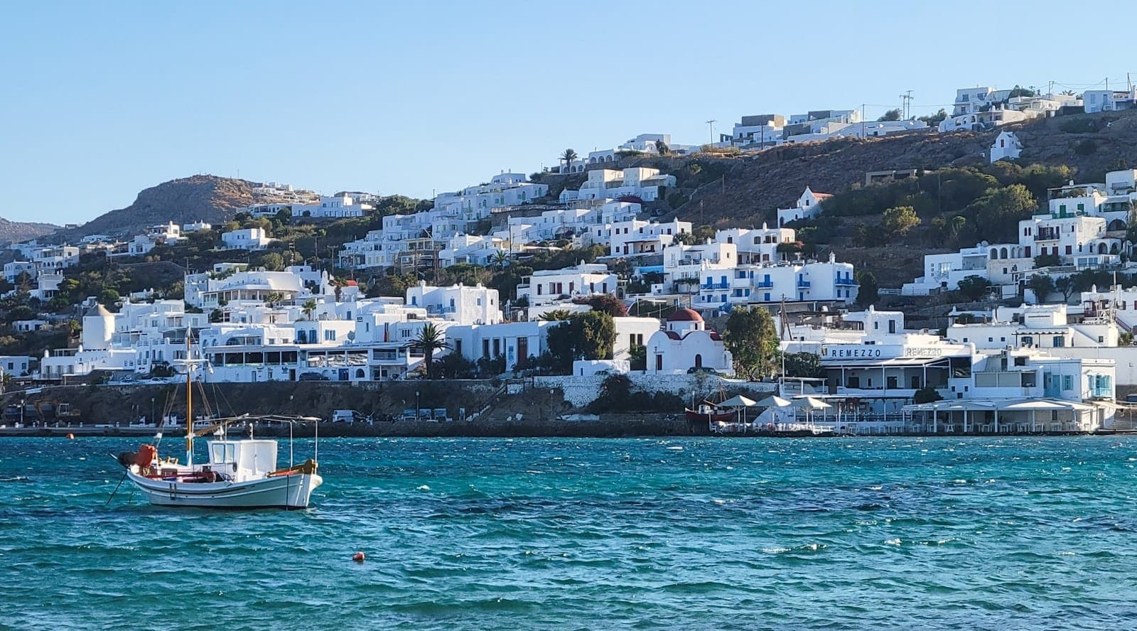 Blick auf die Uferpromenade von Mykonos mit traditionellen weißen Gebäuden und einem Boot auf dem blauen Ägäischen Meer unter klarem Himmel.