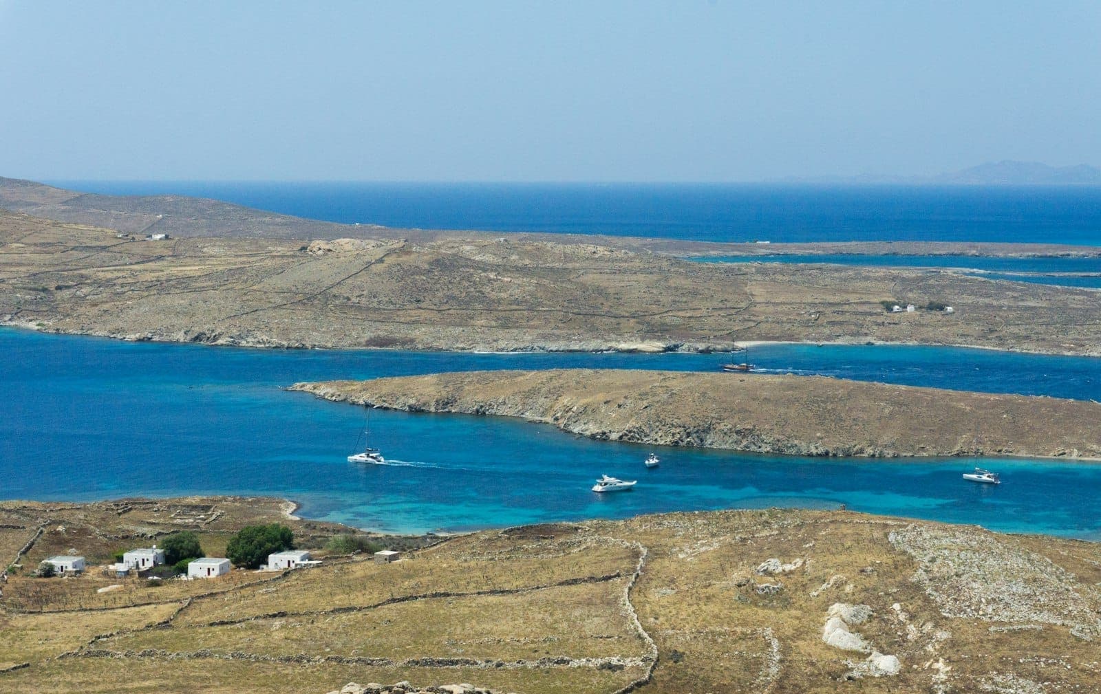Panoramablick auf die karge Insel Delos, umgeben vom tiefblauen Ägäischen Meer, mit kleinen Booten auf dem Weg zwischen Delos und Mykonos unter wolkenlosem Himmel.