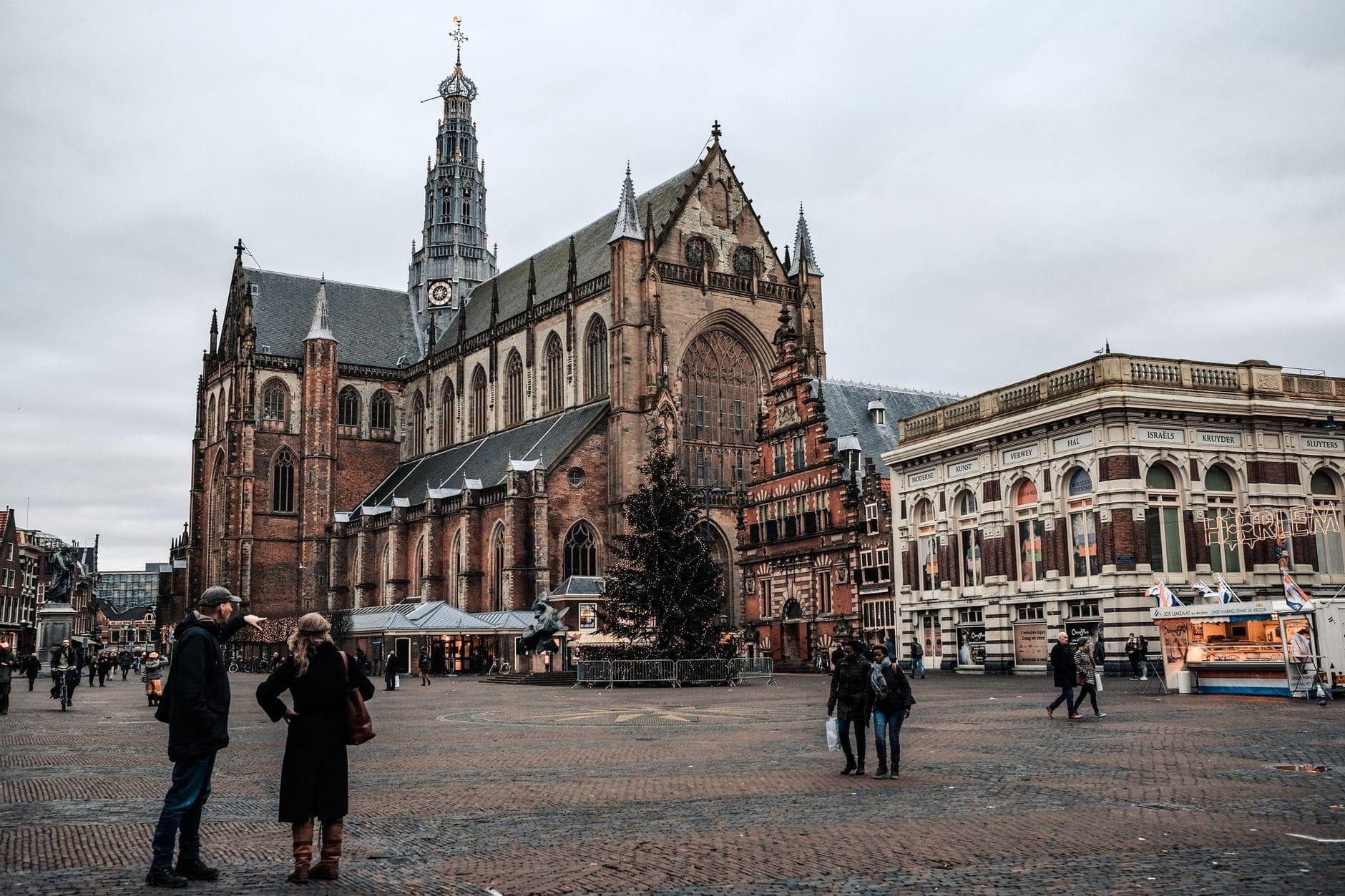 Menschen, die auf einem historischen Platz mit einer großen verzierten Kirche und klassischen Gebäuden unter einem bedeckten Himmel spazieren und sich unterhalten.