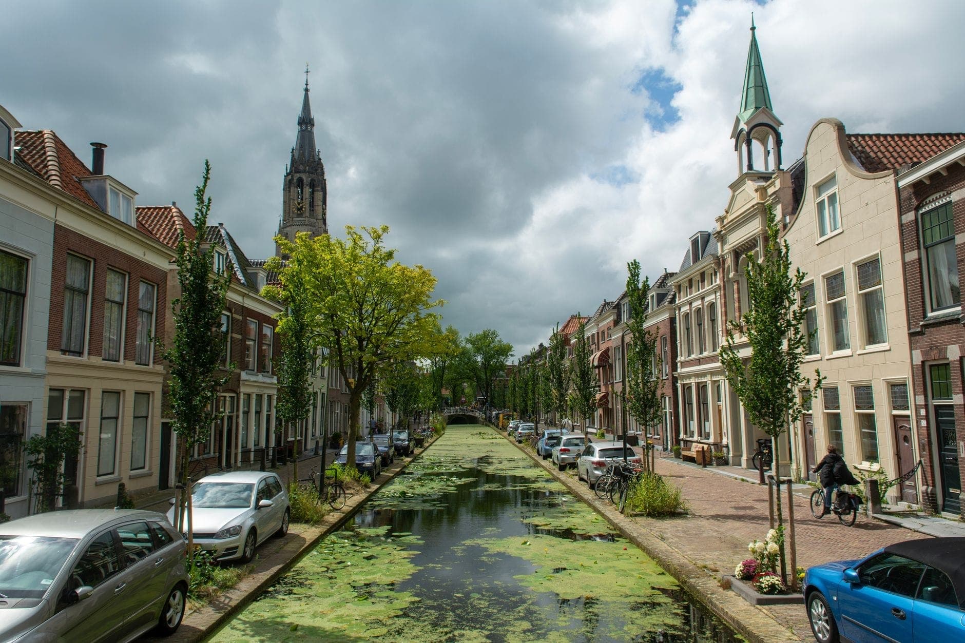 Baumgesäumter Kanal, flankiert von historischen holländischen Häusern, mit einem Kirchturm in der Ferne und am Straßenrand geparkten Autos.
