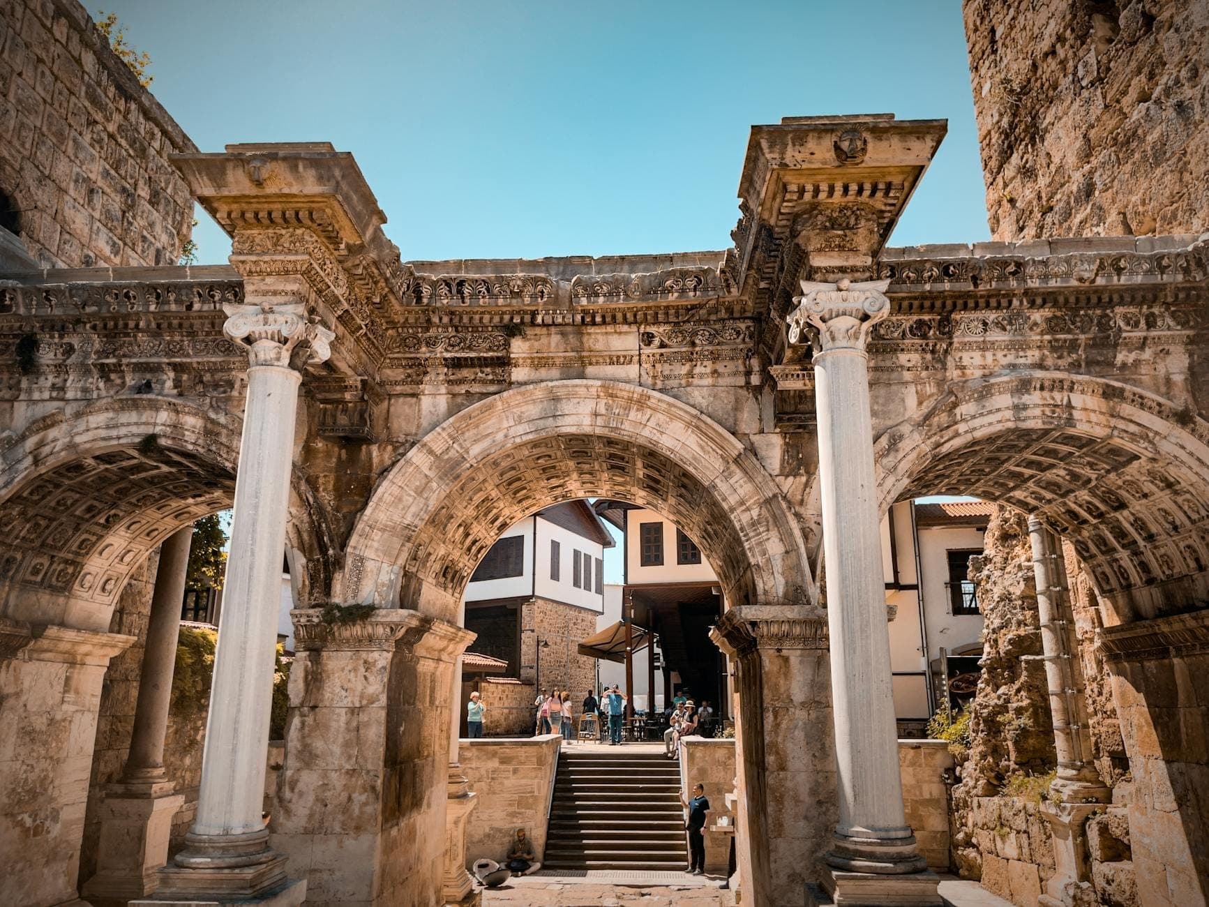 Arche romaine antique avec colonnes en pierre à la Porte d'Hadrien, ouvrant sur une rue de la vieille ville d'Antalya, avec des personnes et des bâtiments historiques visibles.
