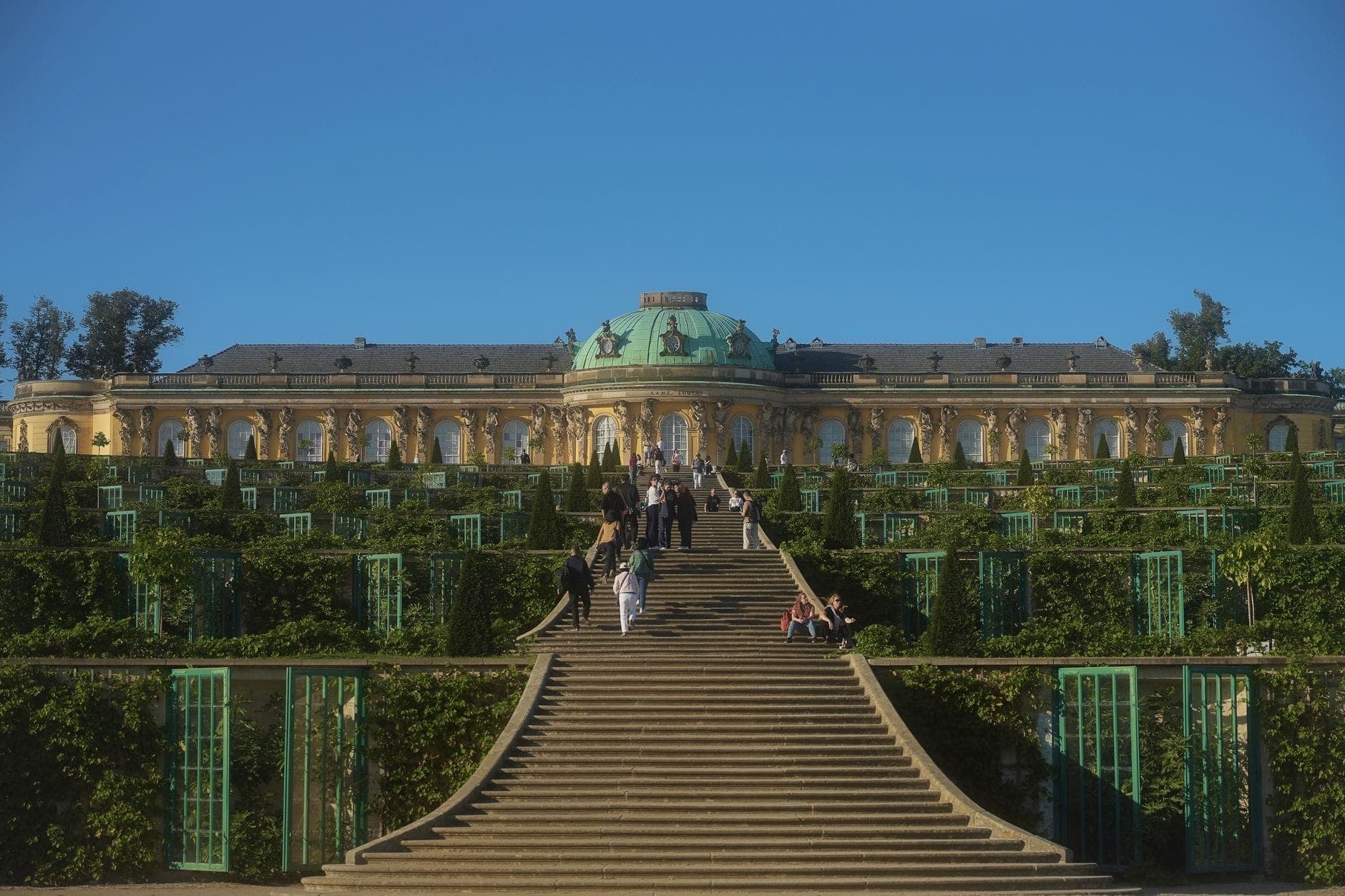 Breite Steintreppe und terrassierte Gärten, die zum prunkvollen Schloss Sanssouci mit grüner Kuppel unter einem klaren blauen Himmel führen.