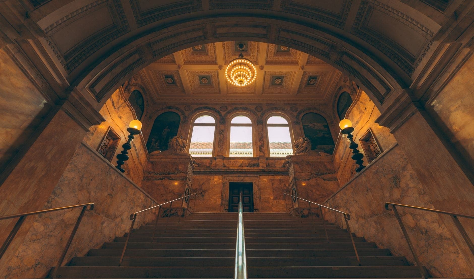Grand escalier en marbre et plafond voûté orné à l'intérieur d'un élégant bâtiment historique, avec des statues et un lustre.