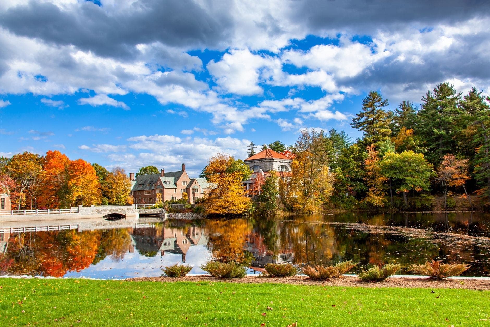 Scène de village avec des bâtiments de style historique, un petit pont en pierre voûté, des arbres d'automne et une eau réfléchissante sous un ciel partiellement nuageux.