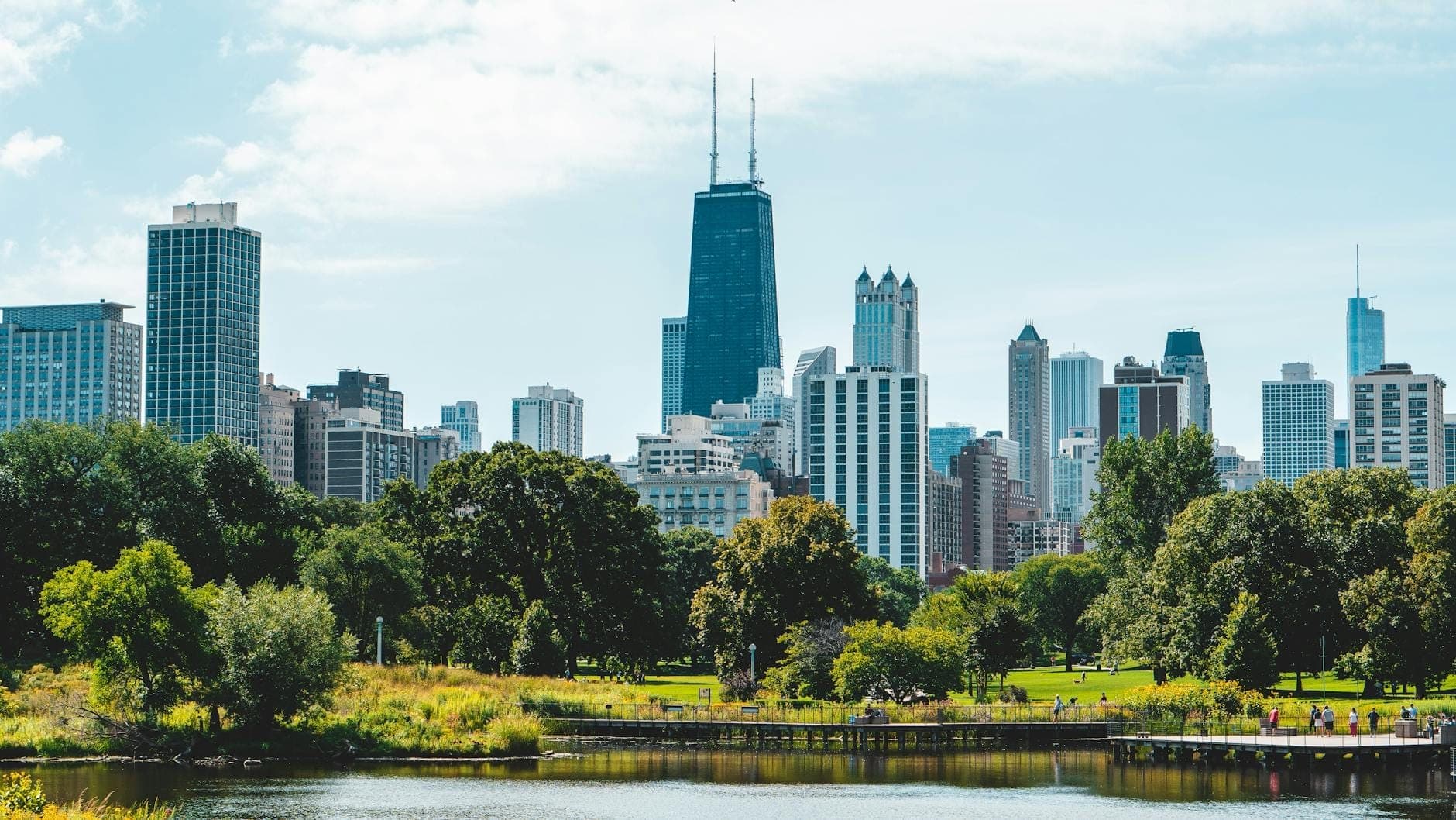 Vista del horizonte de Chicago detrás de un parque verde y un estanque en un día soleado con personas caminando a lo lejos.