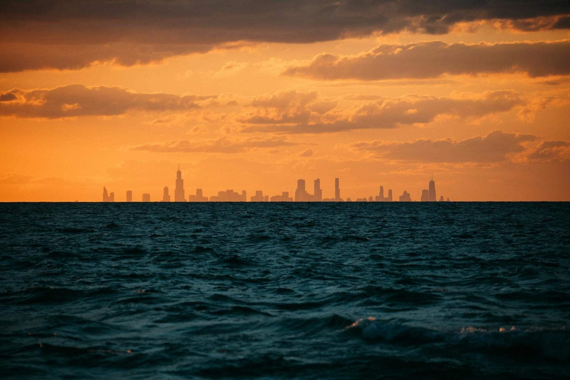 Olas del lago Michigan con el skyline de Chicago silueteado contra un dramático cielo naranja al atardecer.