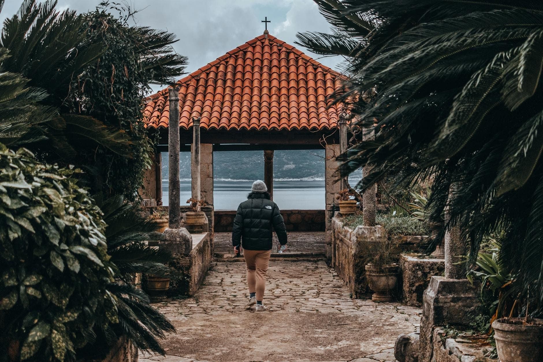 Personne se promenant sur un sentier de jardin luxuriant vers un pavillon avec vue sur la mer, entouré de plantes méditerranéennes près de Dubrovnik.