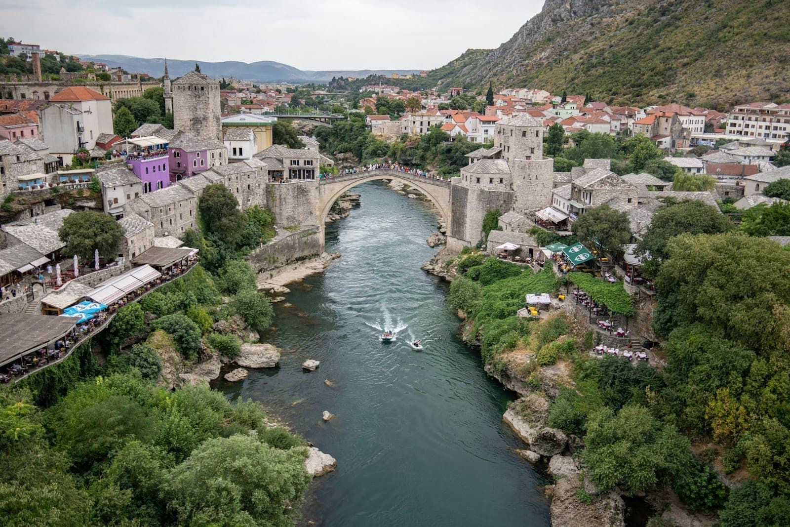 Large vue aérienne de Mostar avec son emblématique pont de pierre enjambant la rivière, entouré de bâtiments traditionnels et de collines verdoyantes.