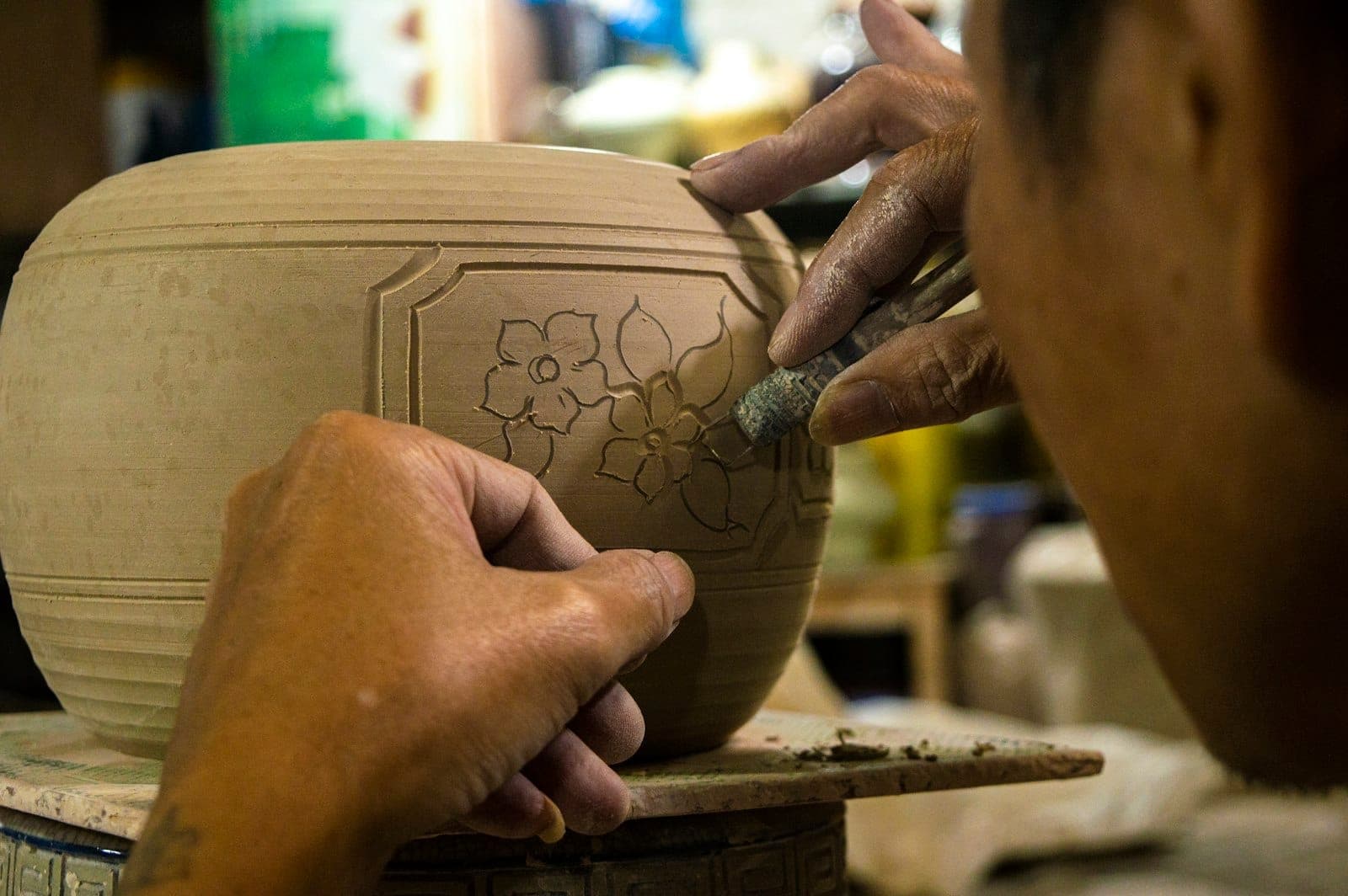Gros plan sur les mains d'un artisan sculptant un motif floral sur un pot en argile dans un atelier de céramique traditionnel.