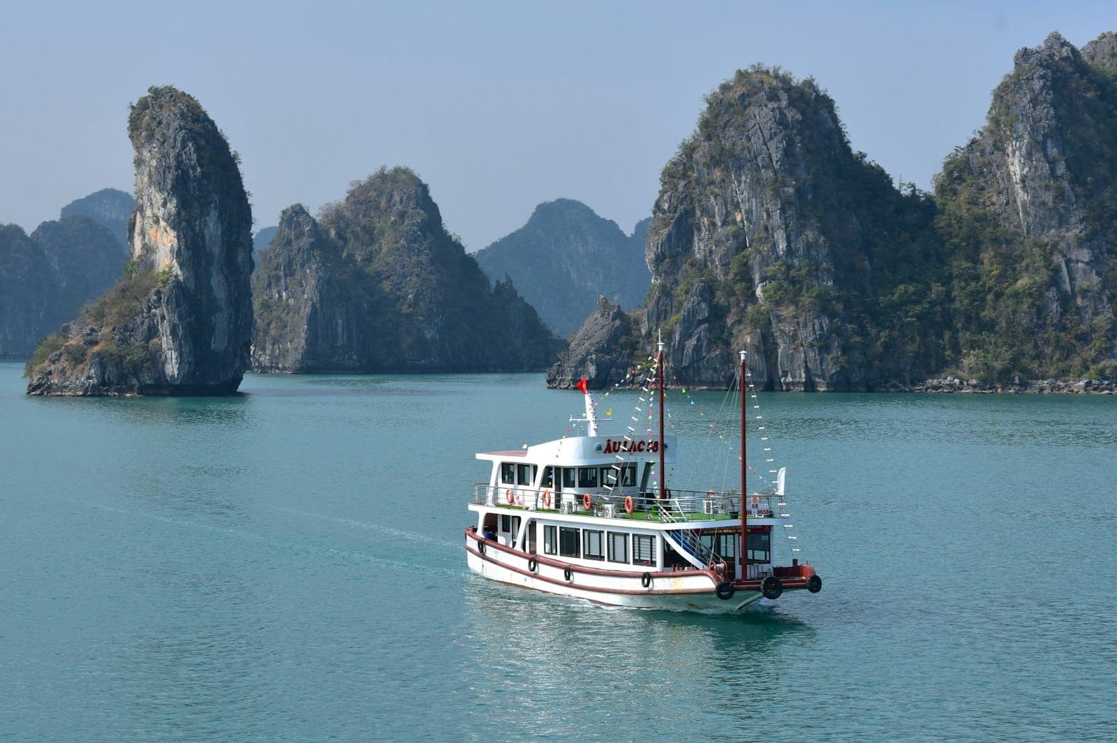 La baie de Ha Long avec un bateau de croisière blanc naviguant parmi des karsts calcaires sur une eau bleue calme sous un ciel dégagé.