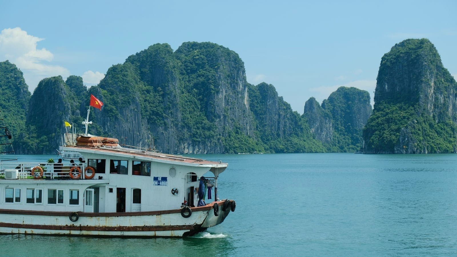 Łódź wycieczkowa płynie po szmaragdowych wodach Ha Long Bay na tle dramatycznych wapiennych krasów pod bezchmurnym, słonecznym niebem.