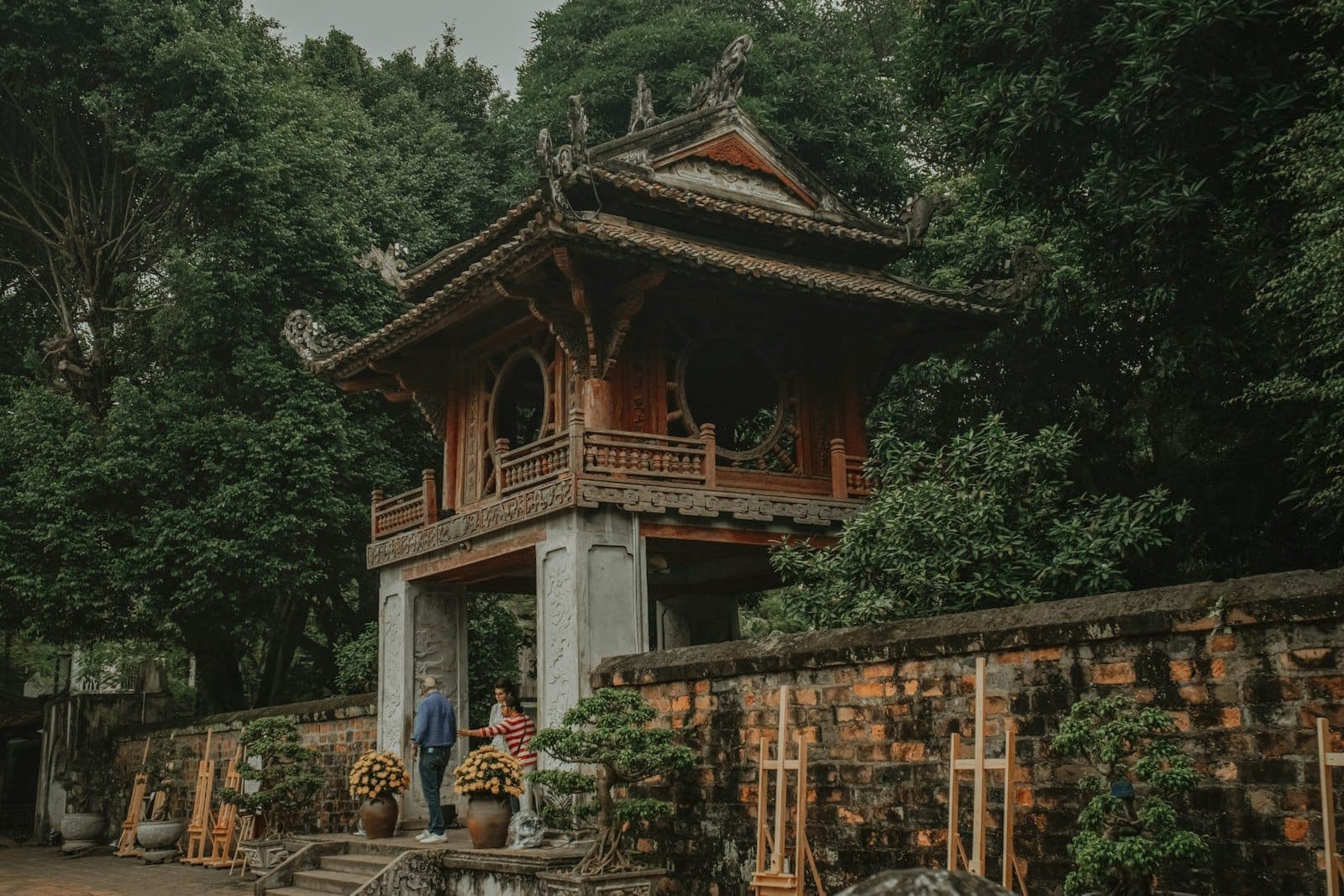 Portail traditionnel d'un temple vietnamien entouré d'arbres luxuriants et de murs en briques, avec deux visiteurs debout sous l'arche.