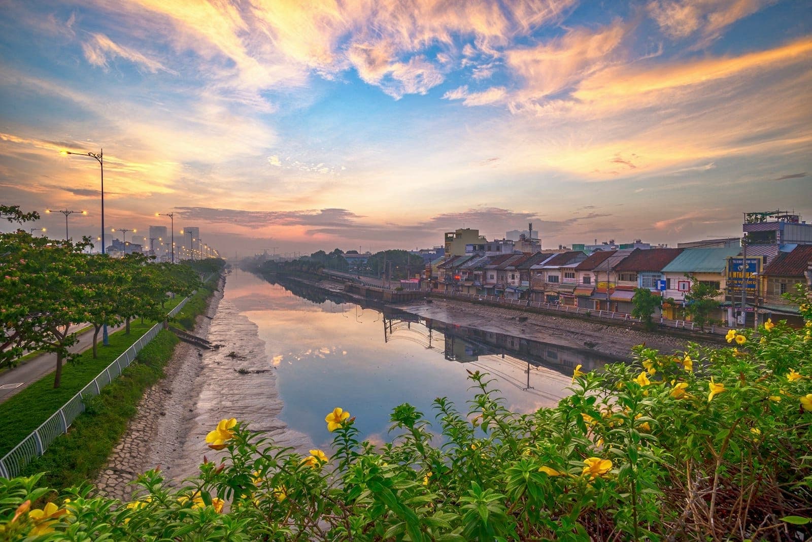 Kanal sungai berwarna-warni saat fajar dengan tanaman berbunga di latar depan dan deretan rumah di tepi air di Ho Chi Minh City, mengundang untuk dijelajahi.