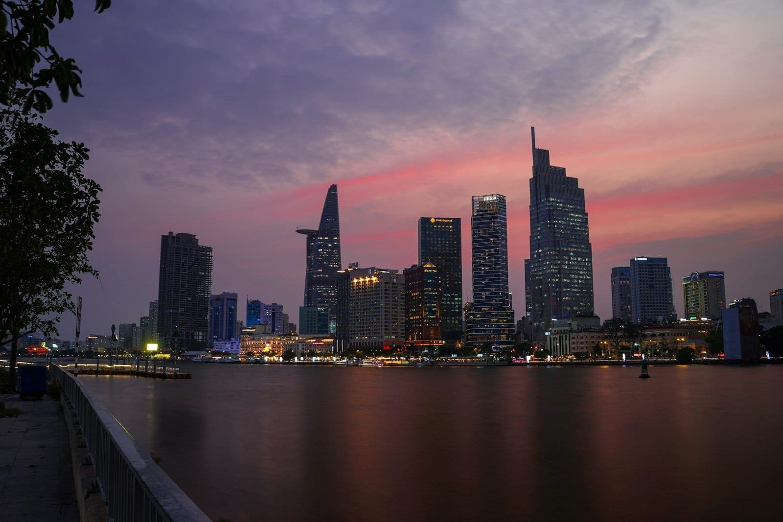 Panorama Ho Chi Minh City o zmierzchu z wieżą Bitexco Tower i innymi nowoczesnymi budynkami odbijającymi się w rzece pod kolorowym niebem.