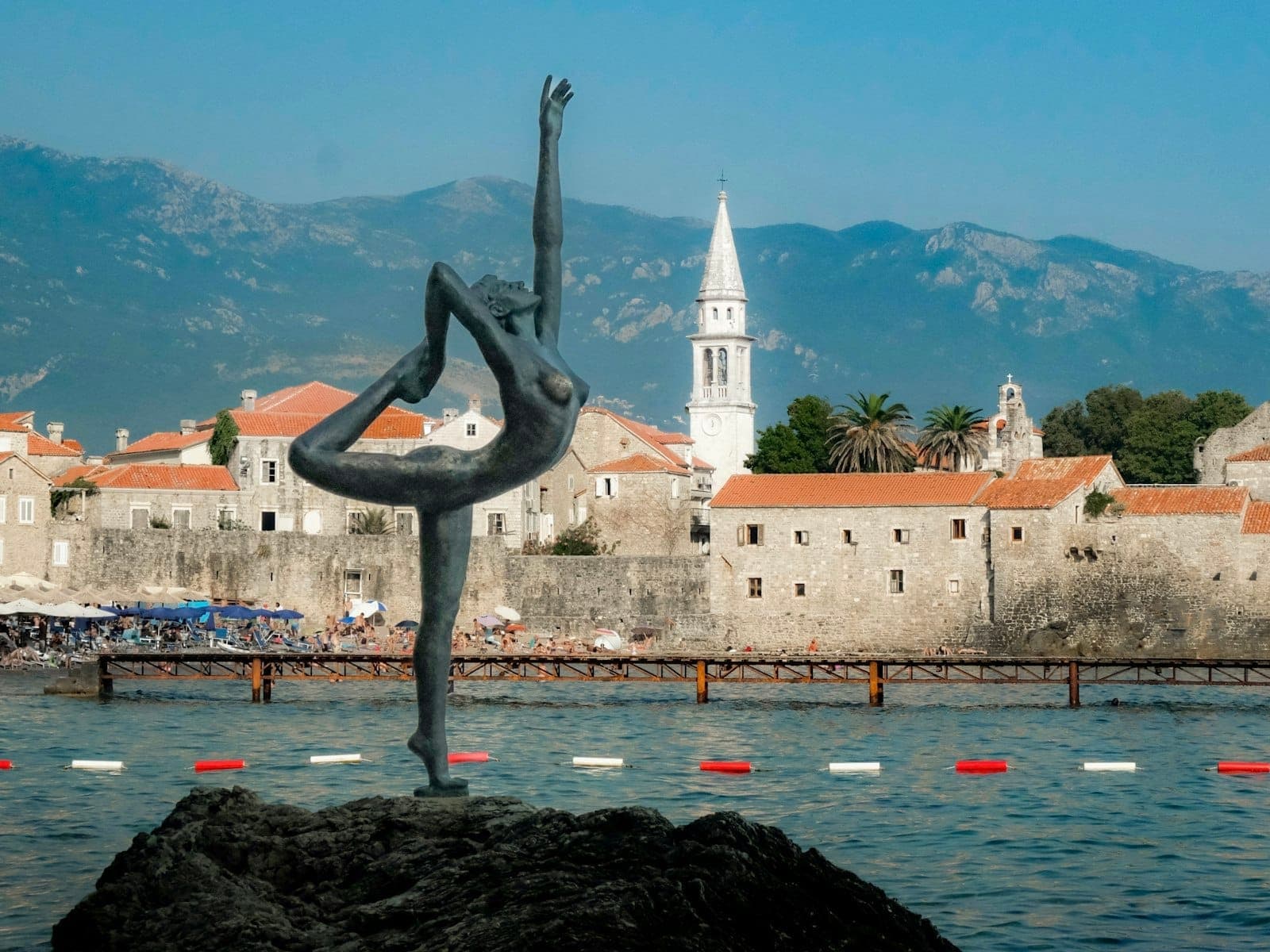 La célèbre statue de ballerine au bord de la mer Adriatique avec les bâtiments en pierre et le clocher de la vieille ville de Budva sur fond de montagnes.