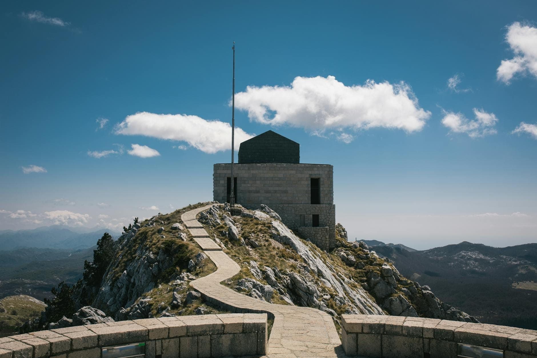 Chemin de pierre menant le long d'une crête rocheuse jusqu'au Mausolée Njegoš dans le Parc National de Lovćen, avec des vues panoramiques sur les montagnes sous un ciel bleu.