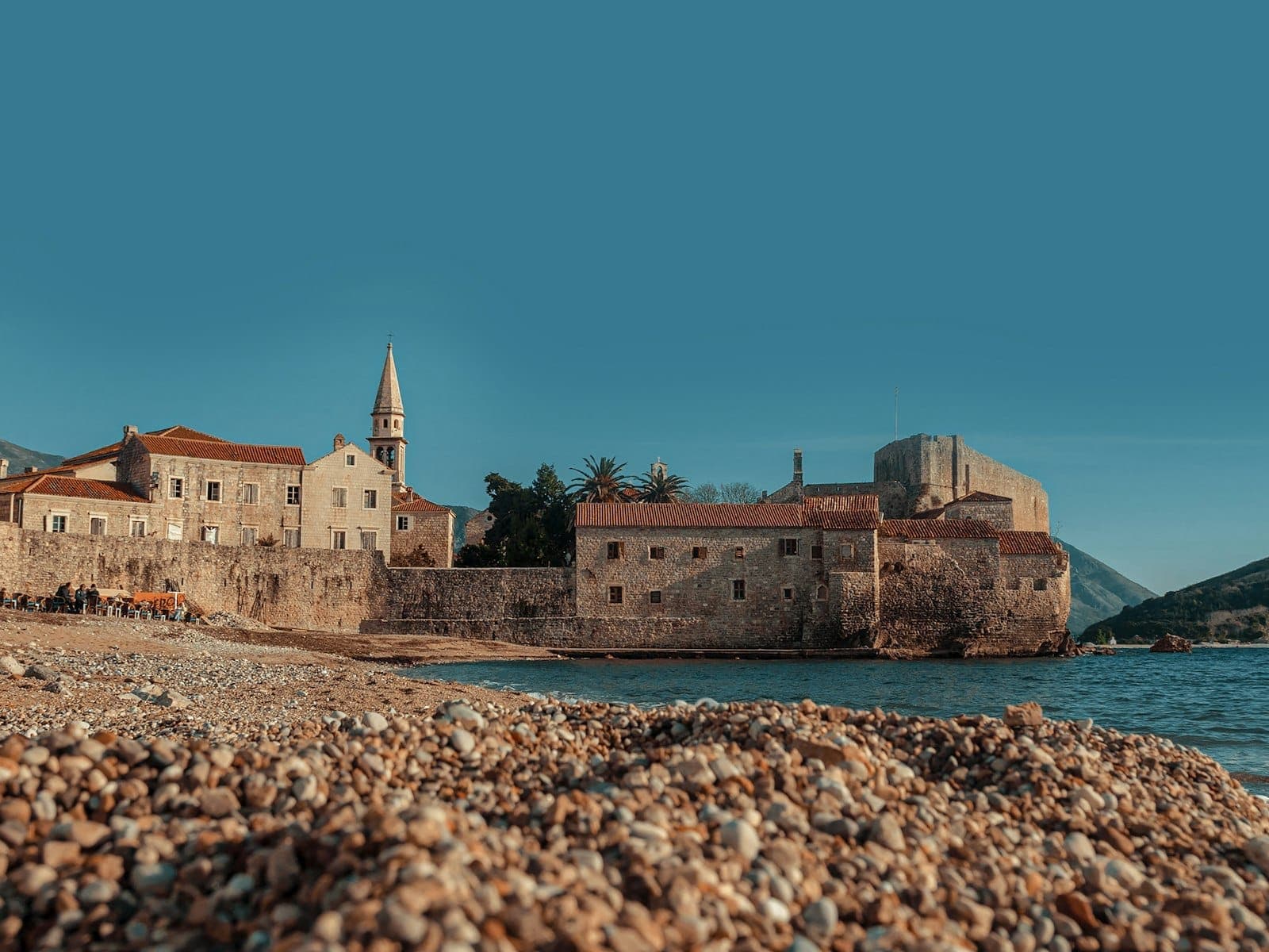 Vieille ville de Budva aux murs de pierre avec clocher et forteresse, vue depuis une plage de galets sous un ciel bleu clair.