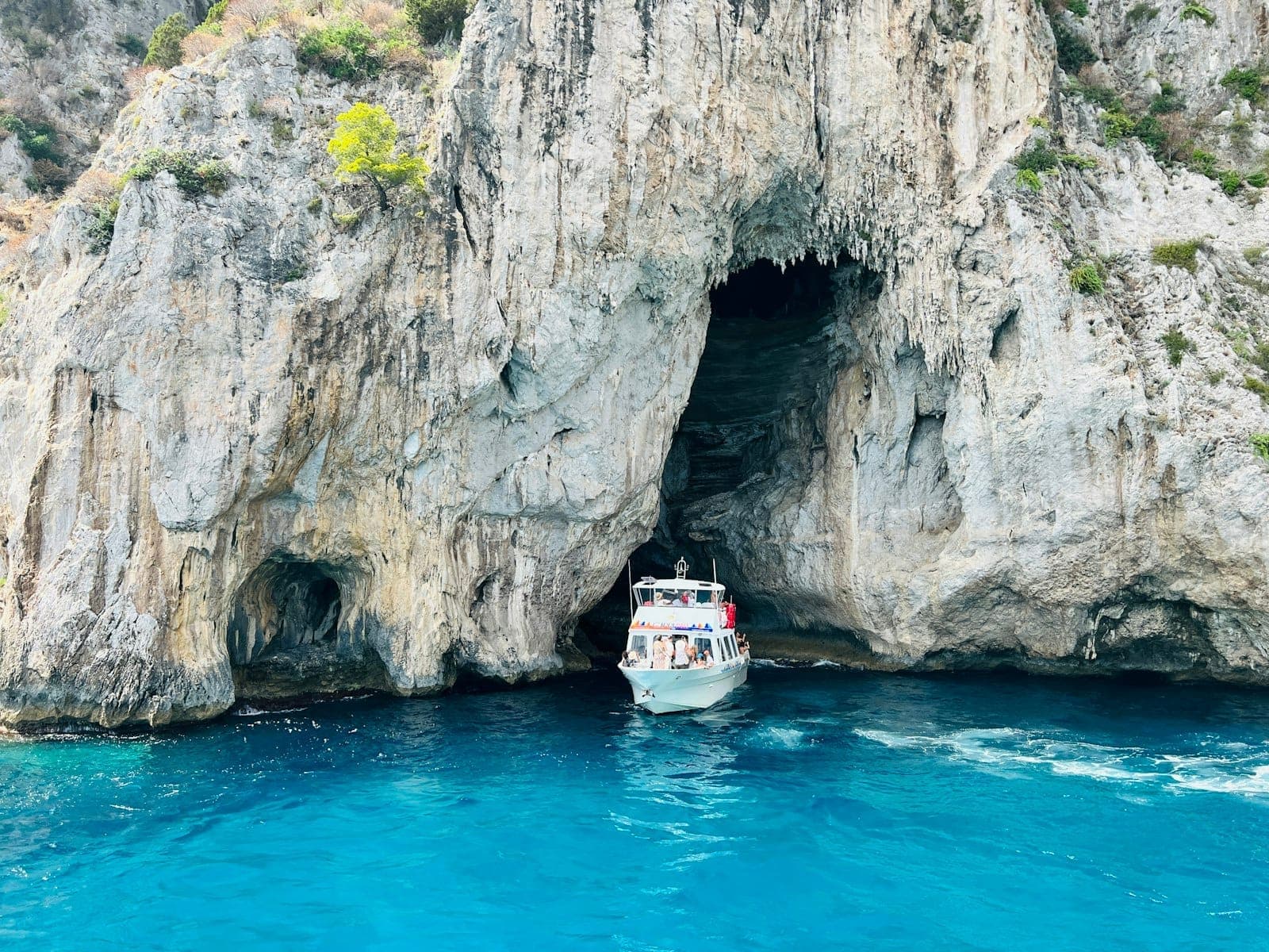Un bateau de visite blanc avec des passagers à bord s'approche de l'entrée d'une grotte marine rocheuse aux eaux d'un bleu vif.