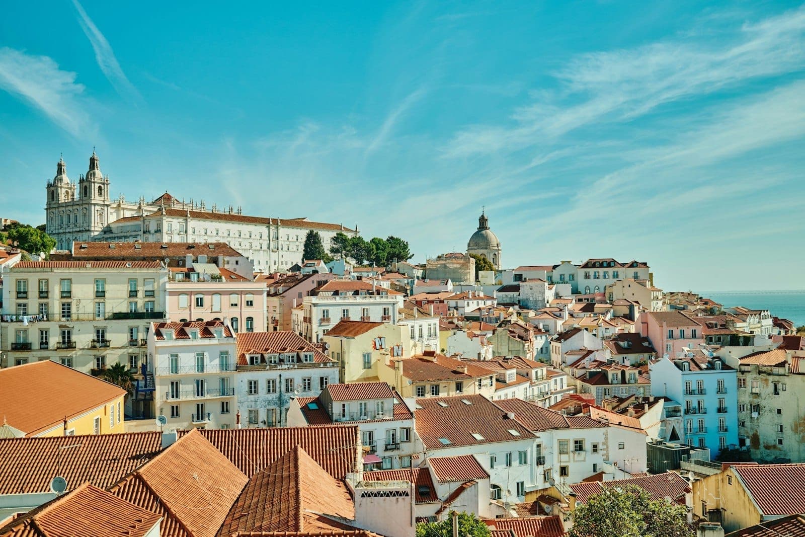 Vista panorâmica e luminosa dos telhados históricos de Lisboa, com prédios brancos, uma igreja com cúpula e céu azul, convidando os visitantes a explorar a cidade e os arredores.