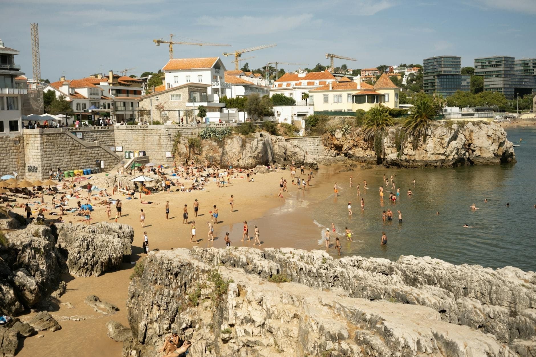 Praia movimentada em Cascais com pessoas a nadar, falésias rochosas e casas históricas, ilustrando o charme costeiro da Linha de Cascais.