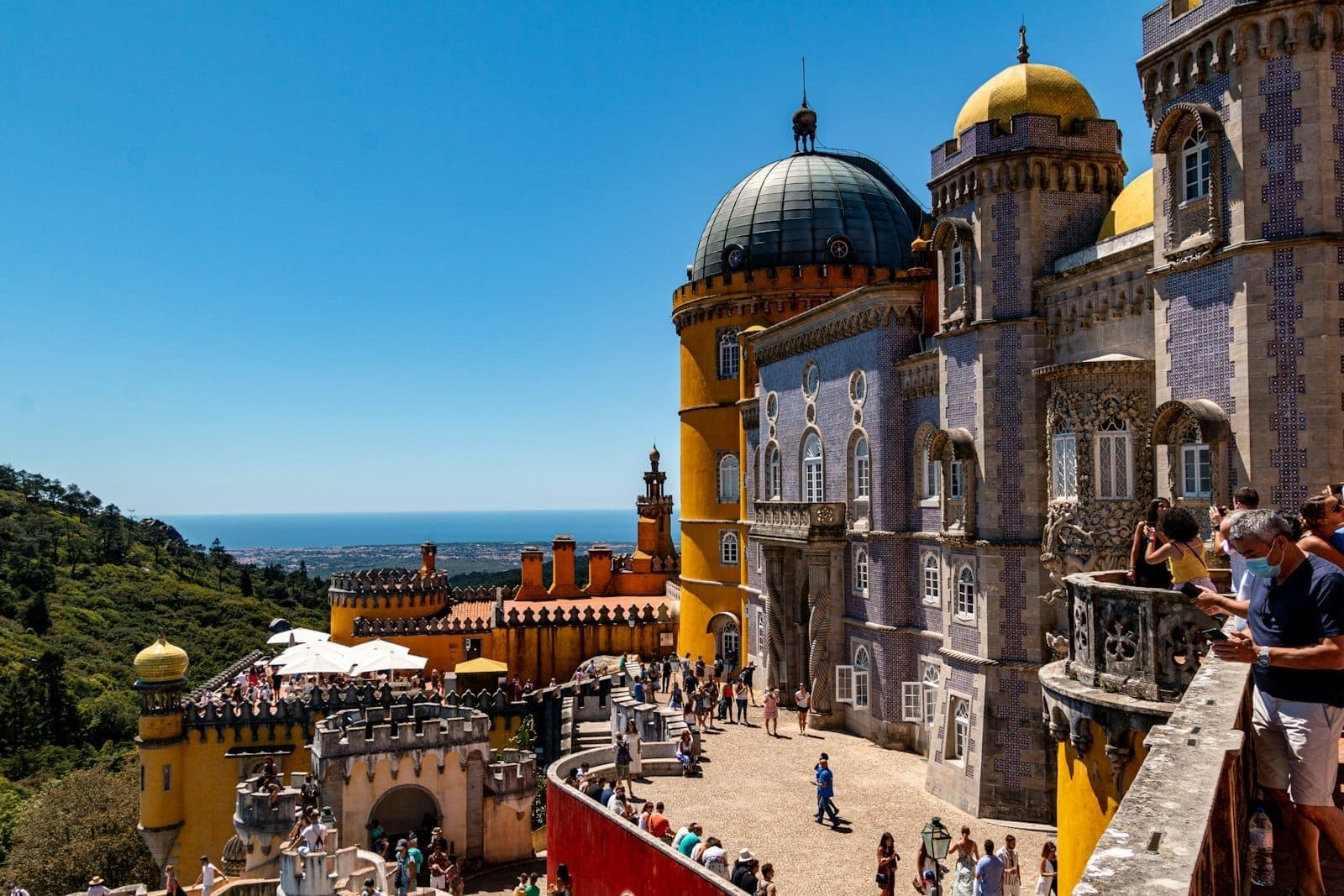 O colorido Palácio da Pena em Sintra com multidões de visitantes, torres decorativas e encostas exuberantes sob um céu azul.