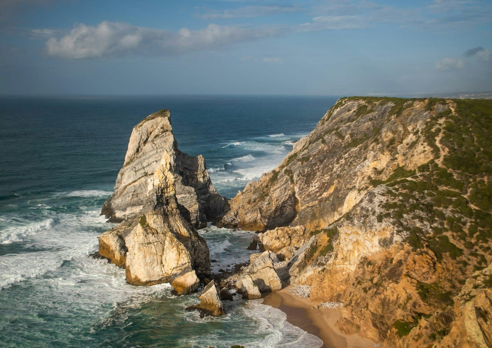 Dramáticos penhascos costeiros e grandes formações rochosas avançando para o Oceano Atlântico com ondas quebrando abaixo e céu azul acima.