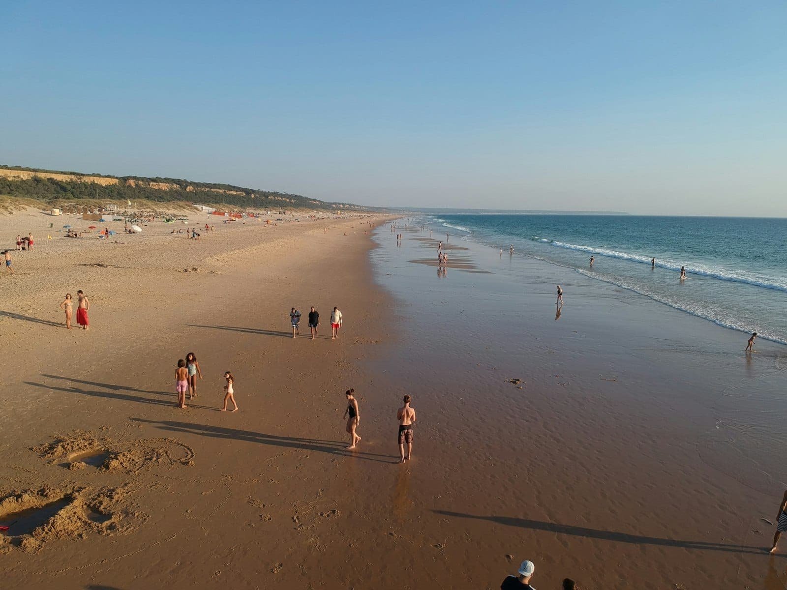 Ampla praia de areia com pessoas caminhando e brincando ao longo da orla sob um céu azul limpo, típica da Costa da Caparica ao sul de Lisboa.