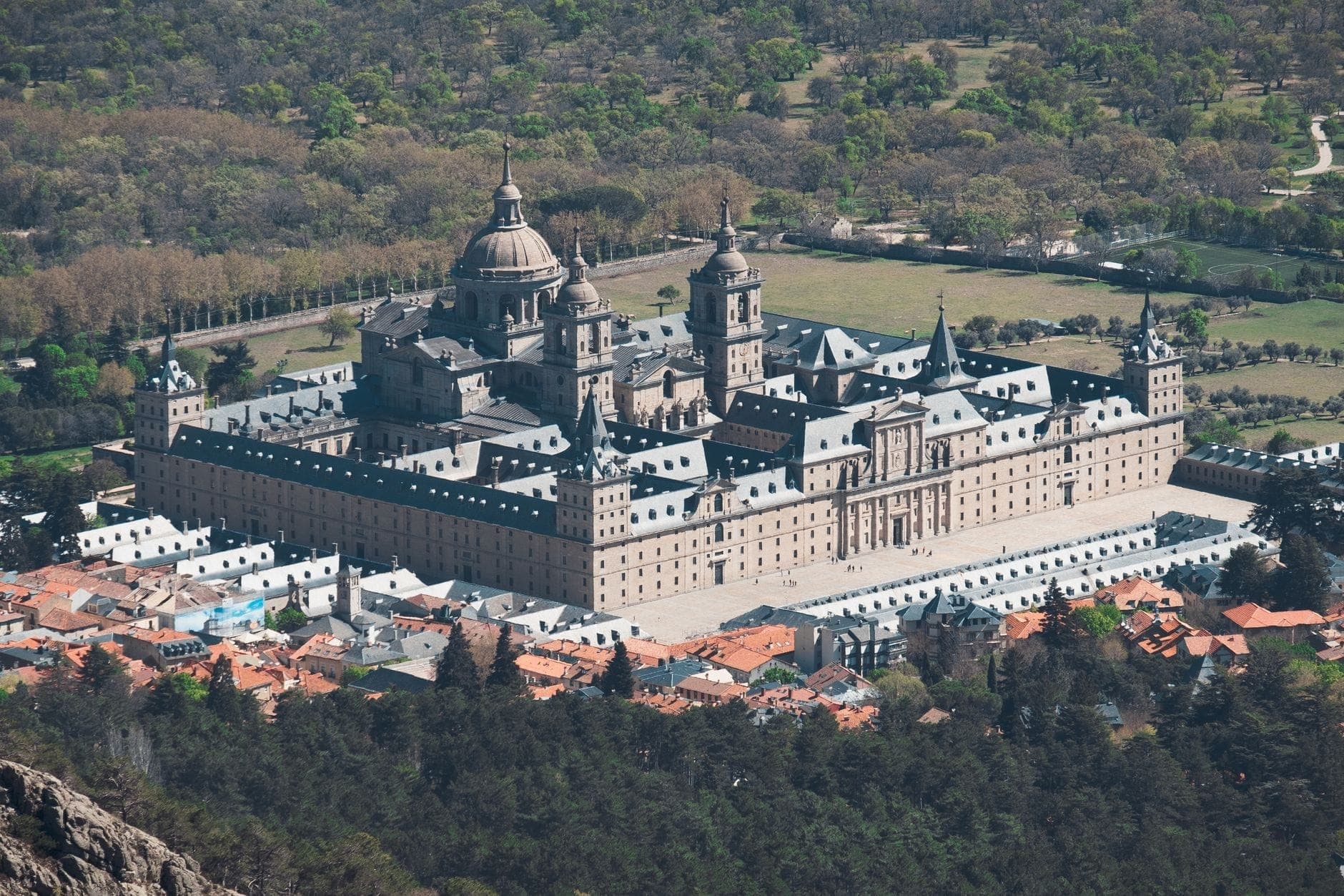 Вид с воздуха на величественный комплекс El Escorial, окружённый зеленью и небольшими крышами, демонстрирующий монументальную архитектуру эпохи Возрождения.