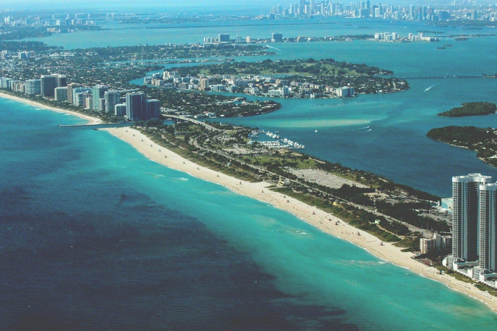 Aerial panoramic view showing the scenic coastline, beaches, and islands near Miami, with turquoise water and city skyline in the distance under a bright blue sky.