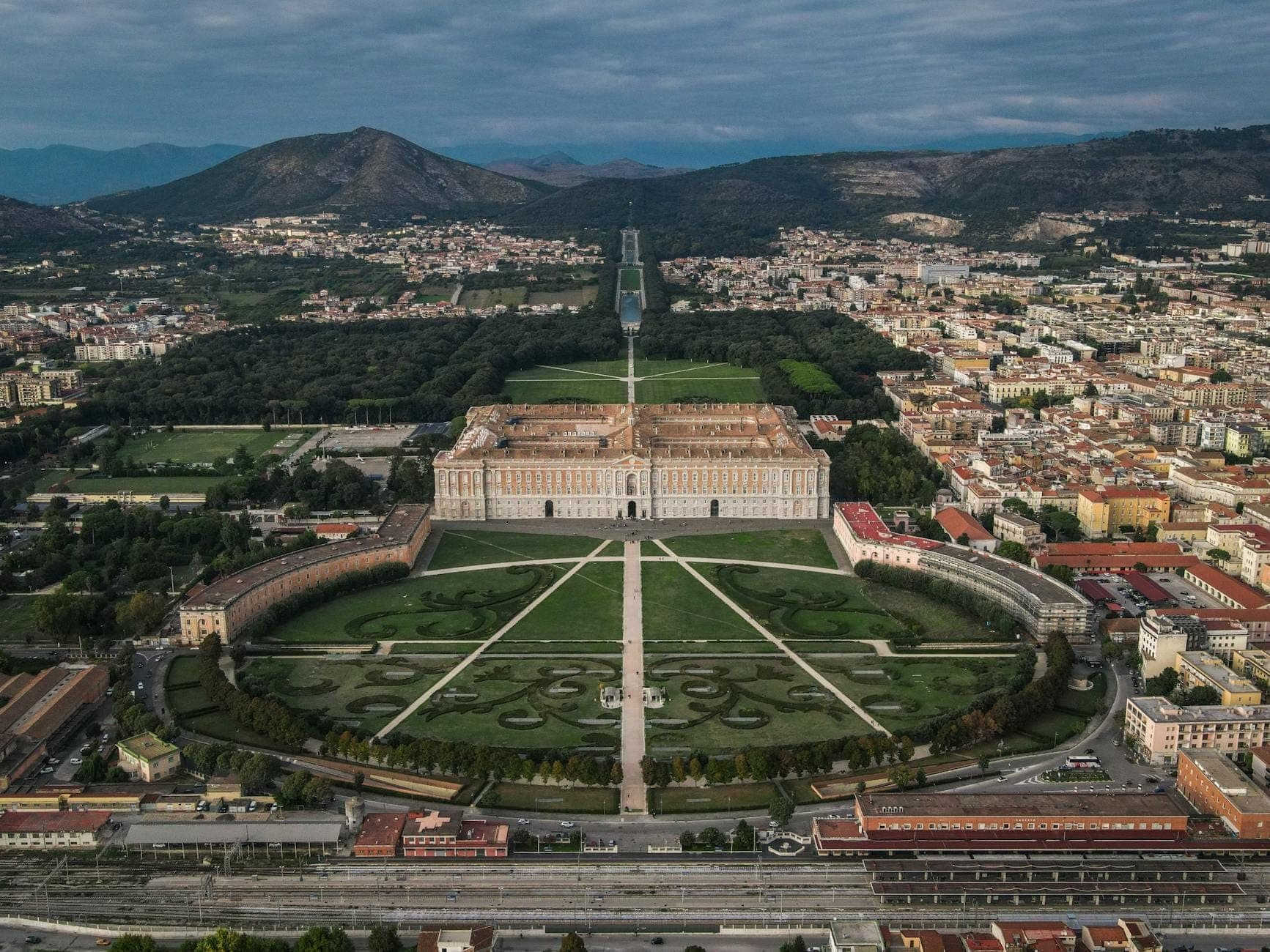 Vista aérea do palácio real da Reggia di Caserta e seus jardins formais rodeados pela cidade e pelas montanhas distantes na Campânia.