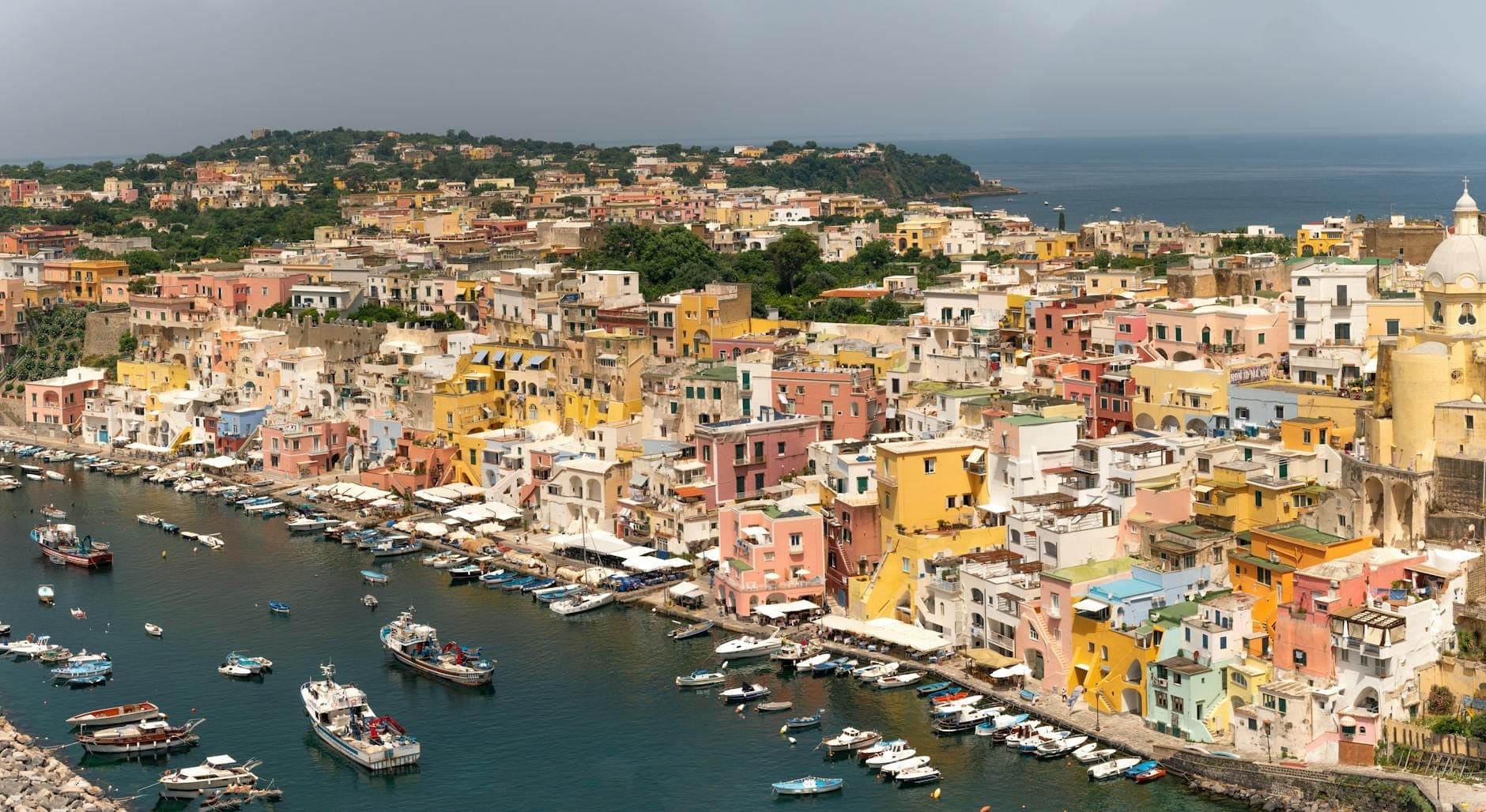 Vista panorâmica de um porto colorido ladeado por edifícios em tons pastel e barcos, com o mar e as colinas verdes ao fundo.