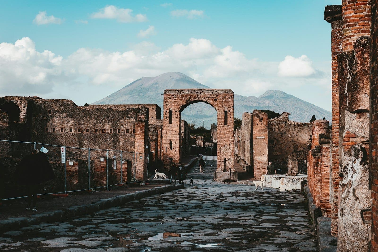 Ruínas de Pompeia com arco de pedra, estrada antiga e o Monte Vesúvio ao fundo sob um céu azul.