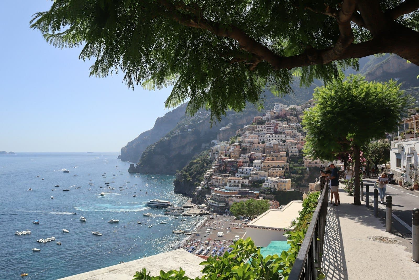 Vista panorâmica de casas coloridas nos penhascos acima do mar em Positano, Costa Amalfitana, com barcos na água e pessoas em um caminho.