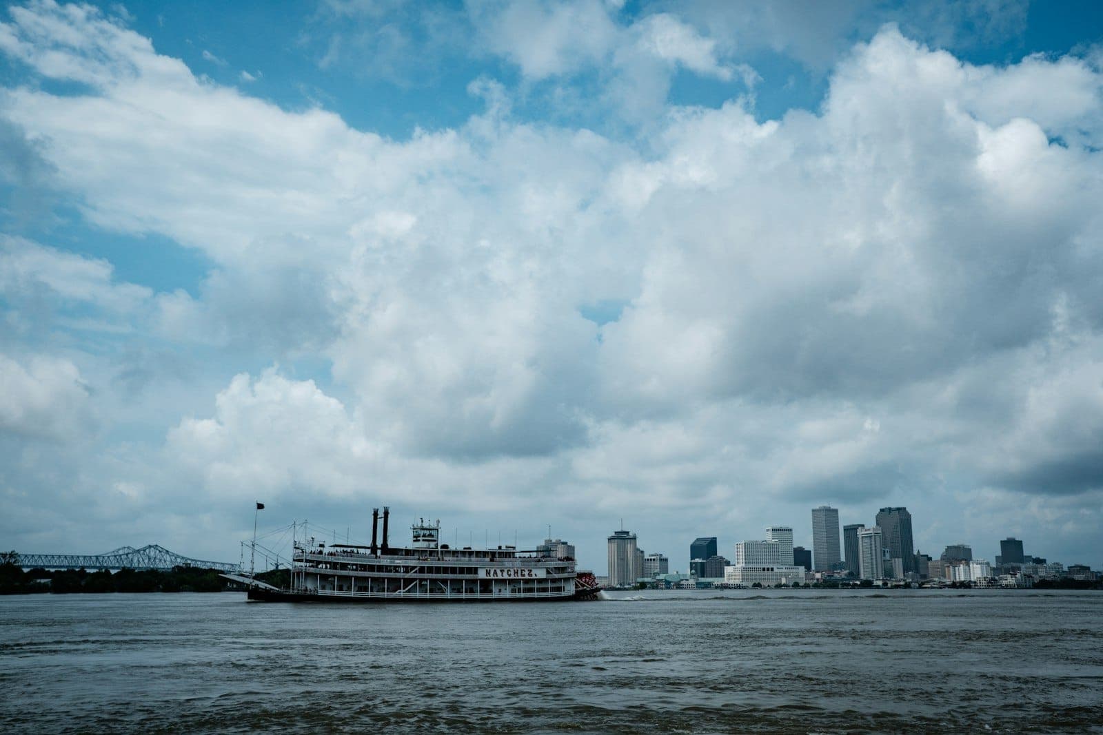 A riverboat travels along the Mississippi River with the New Orleans skyline in the background under a dramatic cloudy sky.