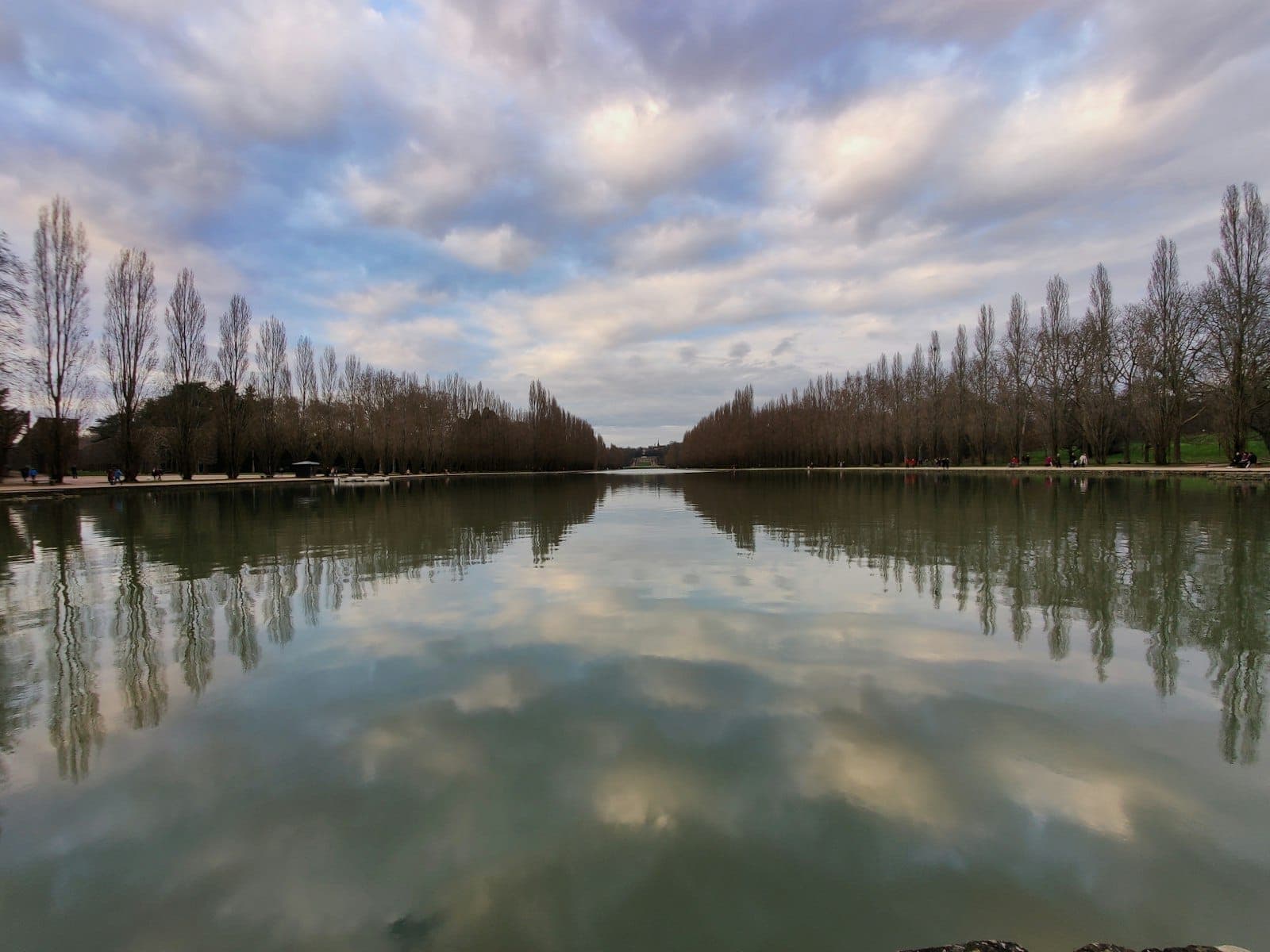 Un long bassin d'eau bordé d'arbres aux reflets symétriques, encadré par des arbres dénudés sous un ciel dramatique, évoquant un grand jardin près de Paris.