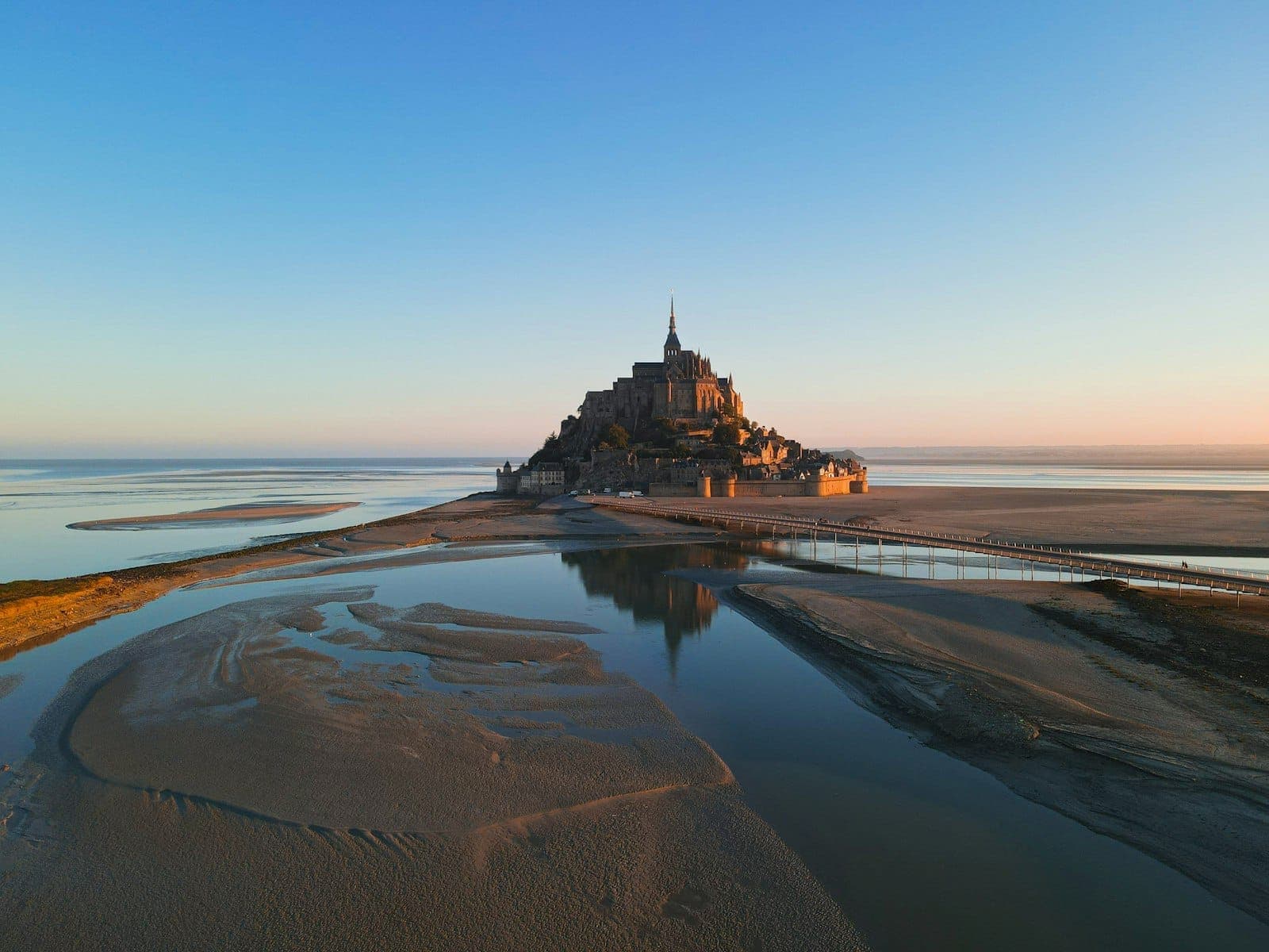Mont Saint-Michel au loin au lever du soleil avec des reflets dans les eaux de marée et un ciel bleu dégagé.