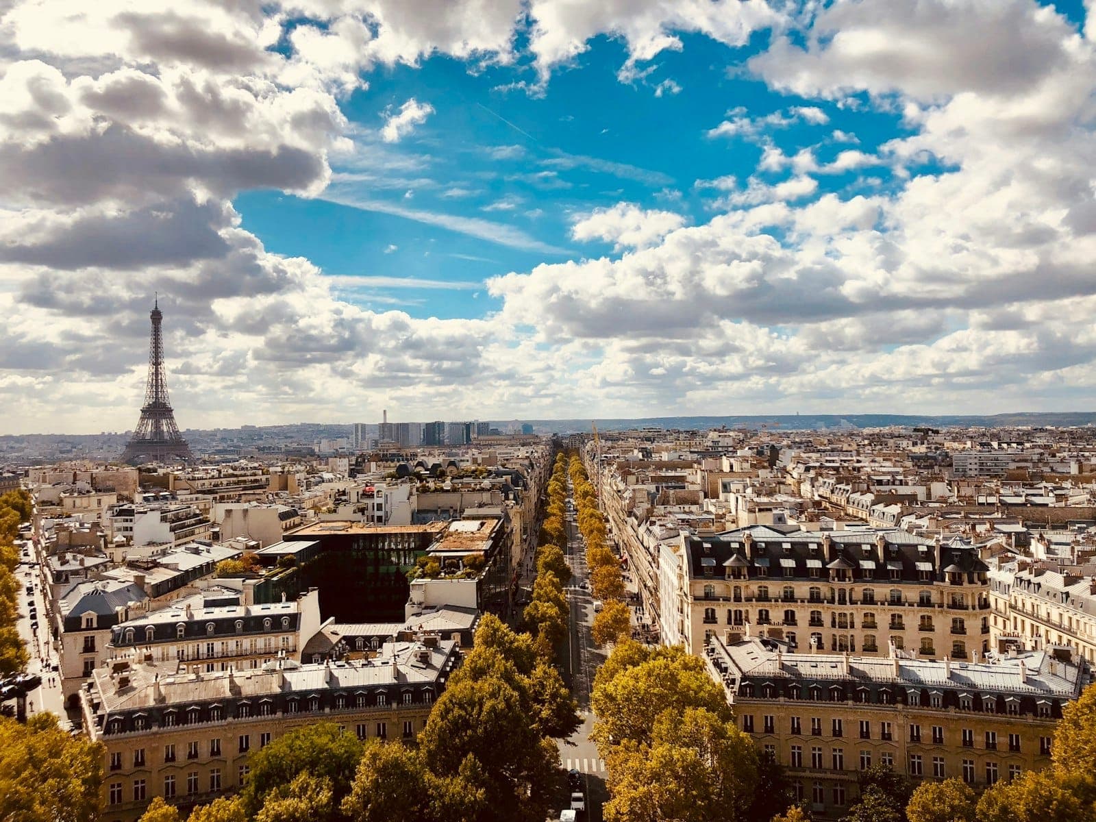 A wide panoramic view of Paris with the Eiffel Tower on the left, tree-lined boulevards, and dramatic blue sky with scattered clouds.