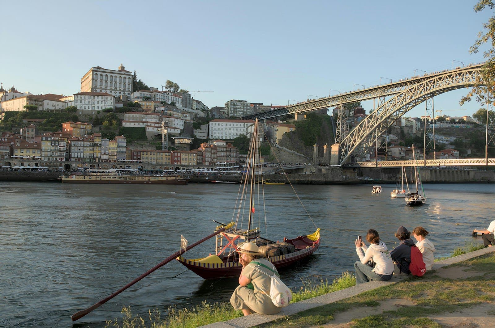 Vila Nova de Gaia'da nehir kenarında dinlenen insanlar, Douro üzerindeki geleneksel tekneler ve Porto'ya bağlanan Dom Luís I Köprüsü