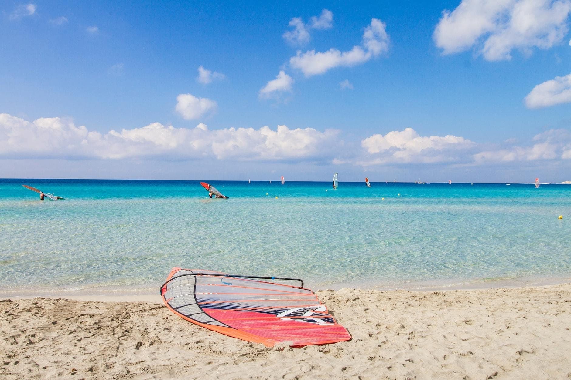 Piaszczysta plaża na południu Rodos z krystalicznie turkusową wodą, deski windsurfingowe i windsurferzy w oddali pod błękitnym niebem.