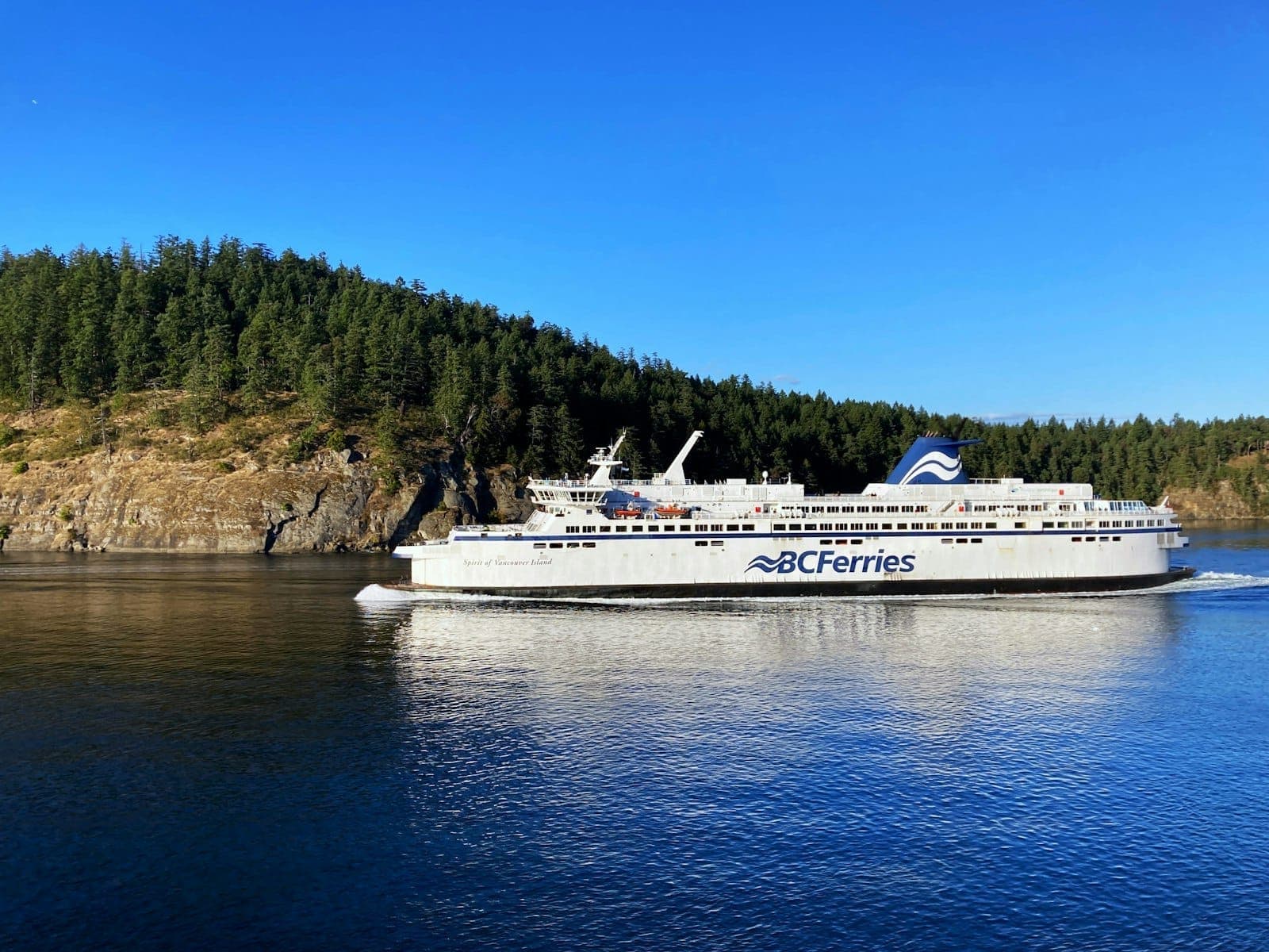 เรือเฟอร์รี่ของ BC Ferries แล่นใกล้เกาะที่ปกคลุมด้วยป่าไม้ภายใต้ท้องฟ้าสีฟ้าแจ่มใส พร้อมชายฝั่งขรุขระและน้ำสีฟ้าเข้มใน Gulf of Georgia