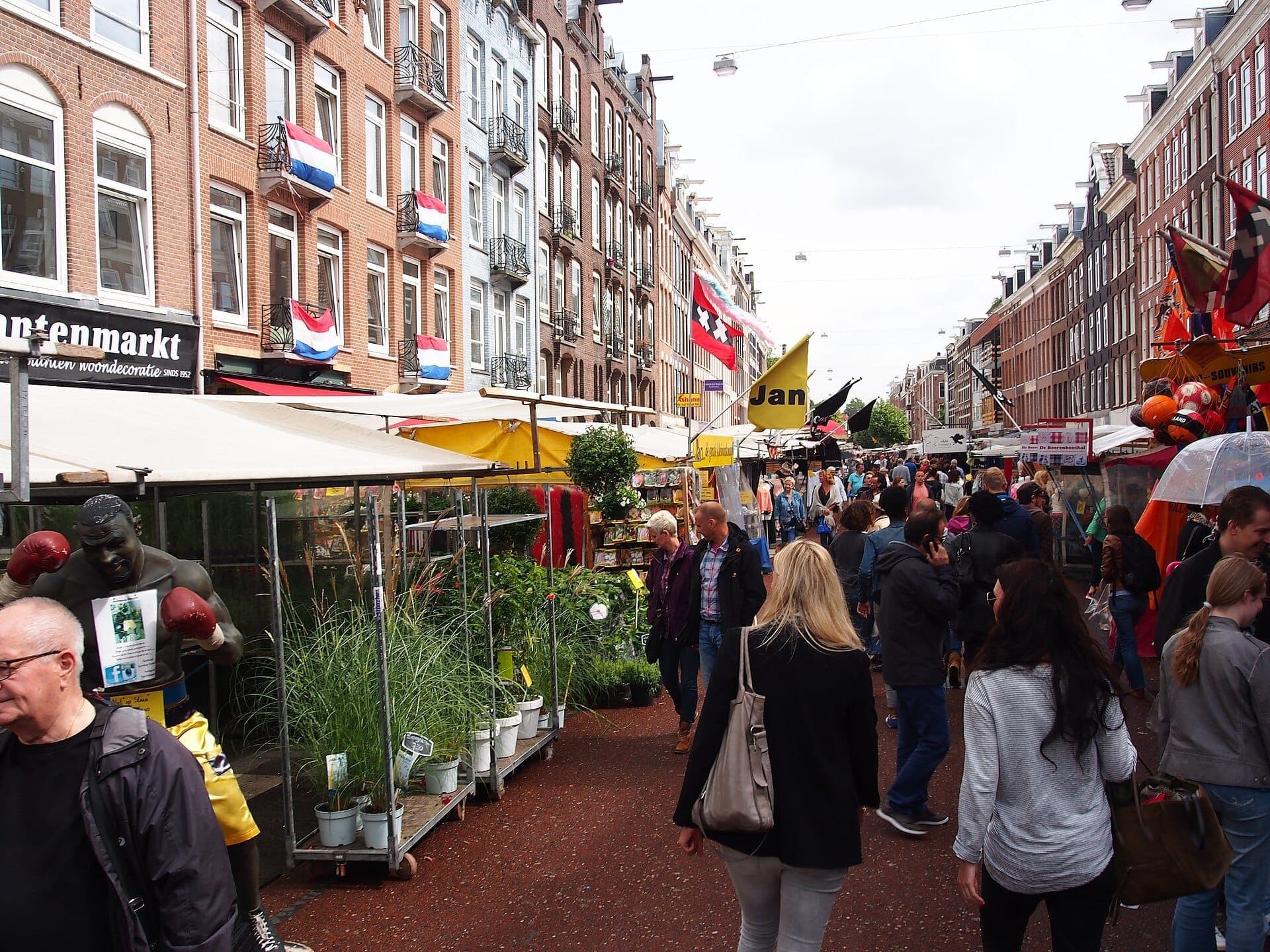 Belebte Straßenszene auf dem Albert Cuyp Markt in De Pijp, mit Backsteingebäuden, niederländischen Flaggen, bunten Marktständen und Menschenmassen beim Einkaufen im Freien.
