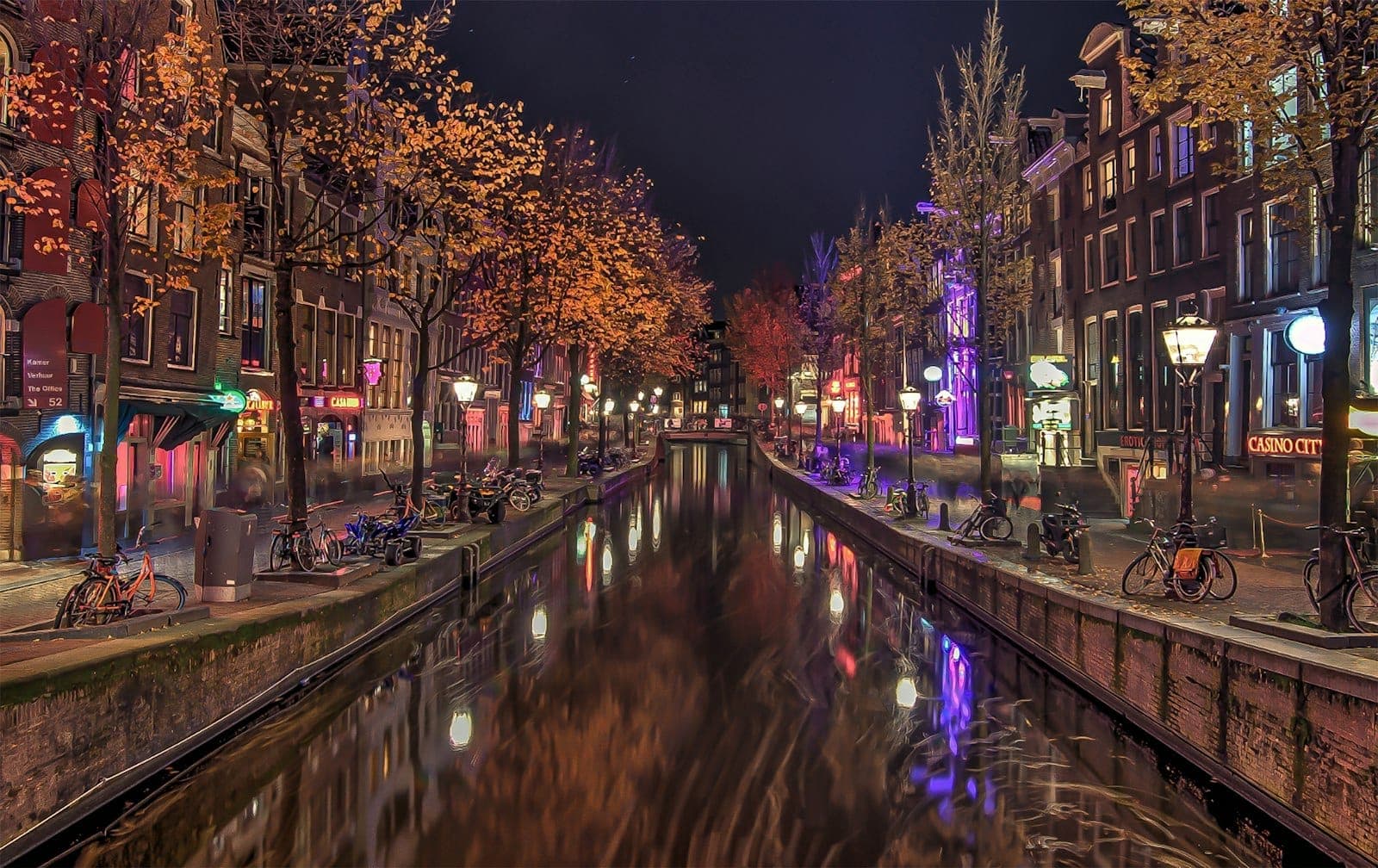 Nächtlicher Blick auf Amsterdams ikonischen De-Wallen-Kanal mit historischen Häusern, leuchtenden Straßenlaternen, Fahrrädern und farbenfrohen Spiegelungen – die lebhafte Atmosphäre des Rotlichtviertels ist spürbar.