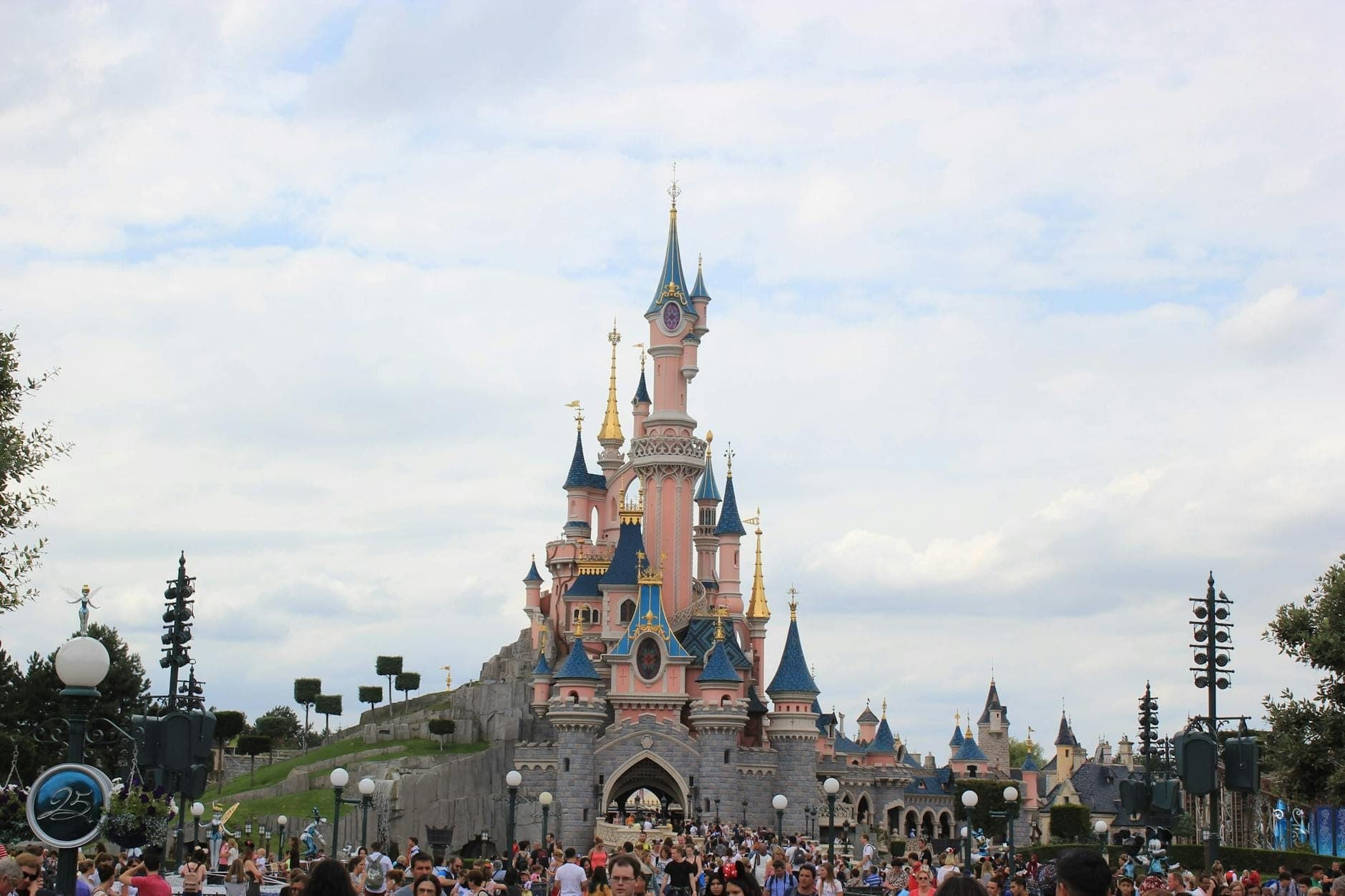 Large crowd in front of the iconic pink Sleeping Beauty Castle at Disneyland Paris under a bright, cloudy sky, capturing excitement and the park’s main landmark.