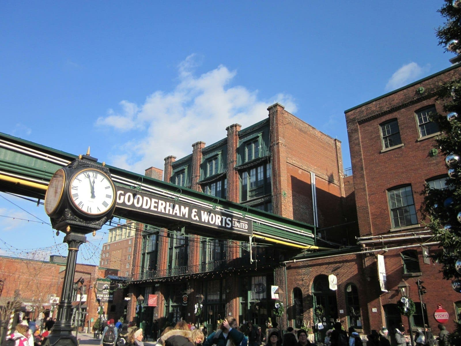 Un'affollata area pedonale del Distillery Historic District con edifici industriali vittoriani in mattoni rossi, l'insegna Gooderham & Worts e un orologio decorativo sotto un cielo soleggiato.