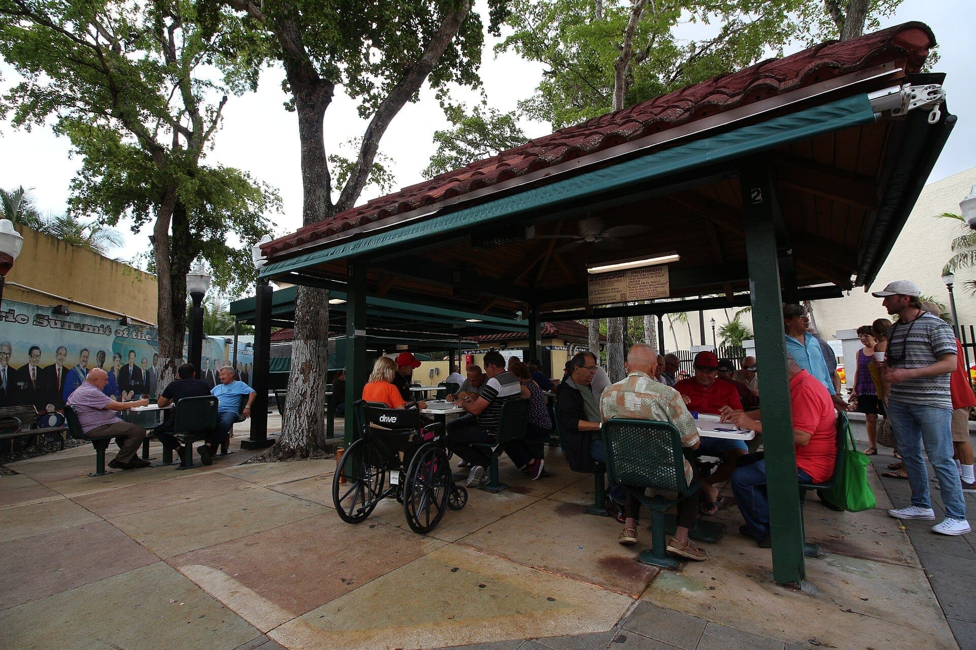 People gather under a covered pavilion at Domino Park, playing dominoes and talking, with trees and a colorful mural in the background.