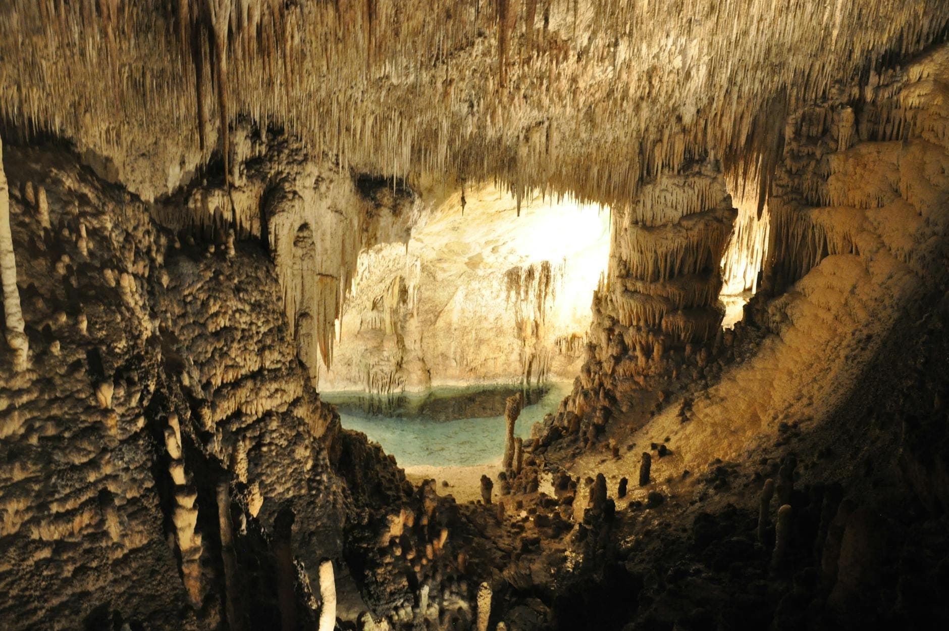 Vista panoramica all'interno di una grotta illuminata con imponenti stalattiti che scendono dal soffitto e un tranquillo lago sotterraneo azzurro sullo sfondo.