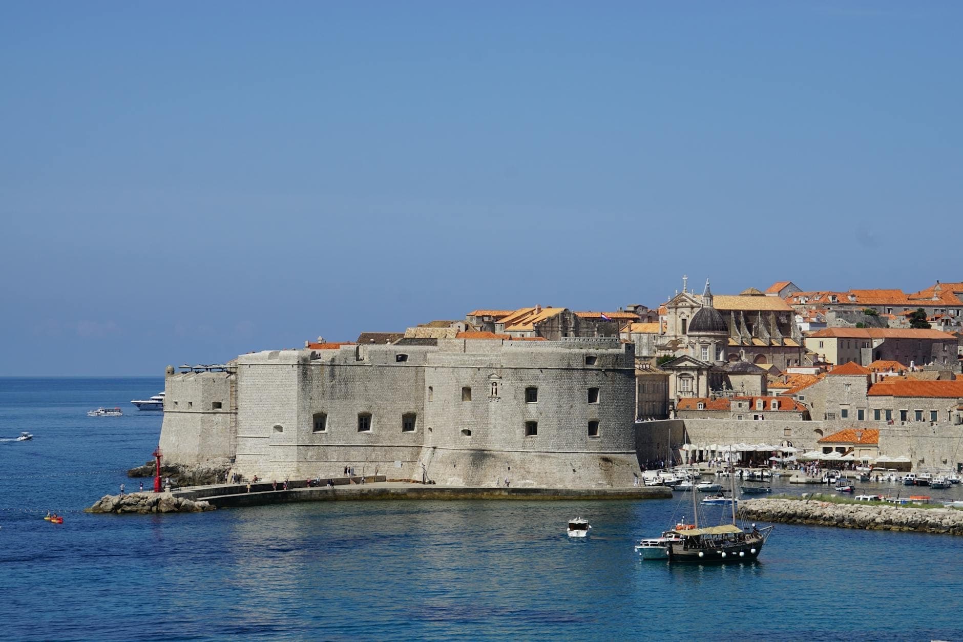 Blick auf die Festung St. Johannes in Dubrovnik am Hafenrand, umgeben von klarem blauem Adriawasser und den orangefarbenen Dächern der Altstadt unter wolkenlosem Himmel.