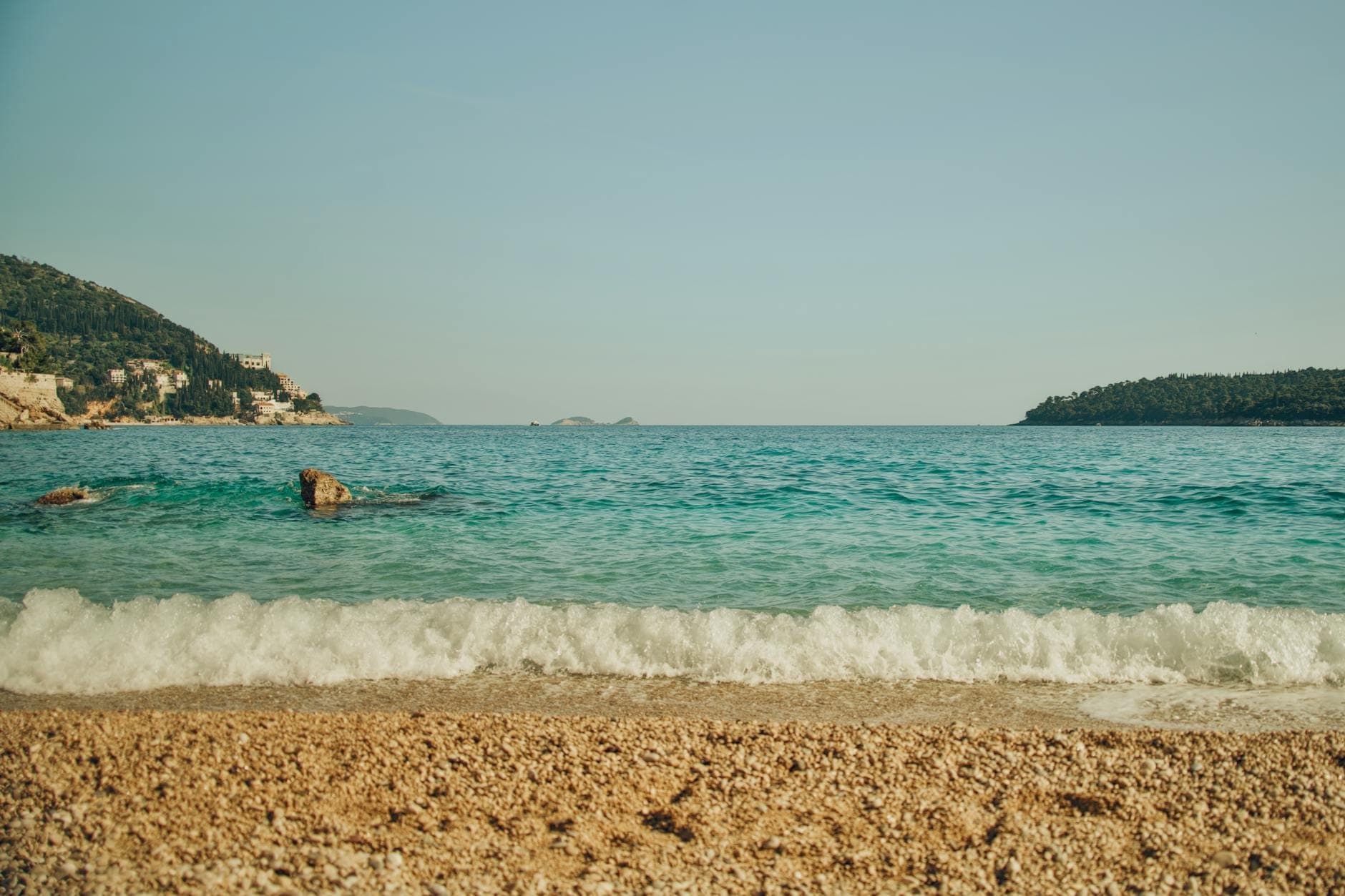 Галечный пляж с бирюзовой водой, спокойными волнами и видом на зелёные холмы и острова вблизи Дубровника в ясный день.